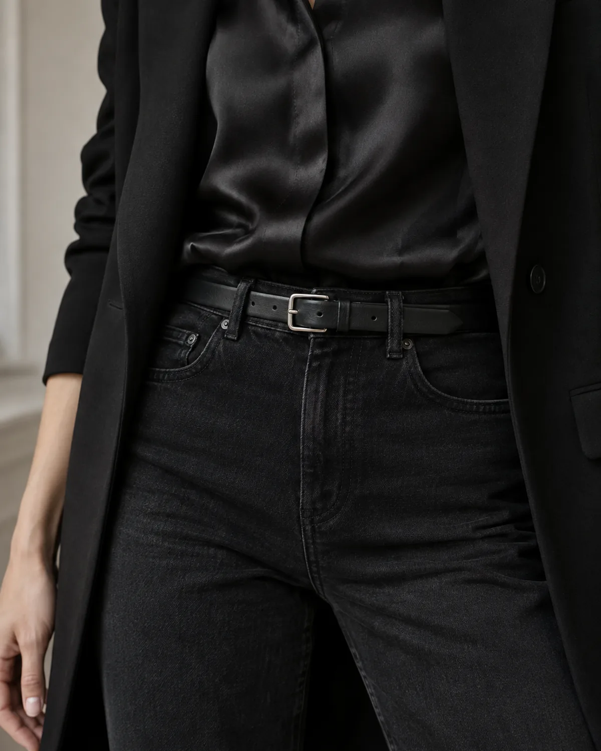 Close crop on the waist area showing a black silk blouse tucked into black straight-leg jeans with a thin black leather belt, under an open black longline blazer
