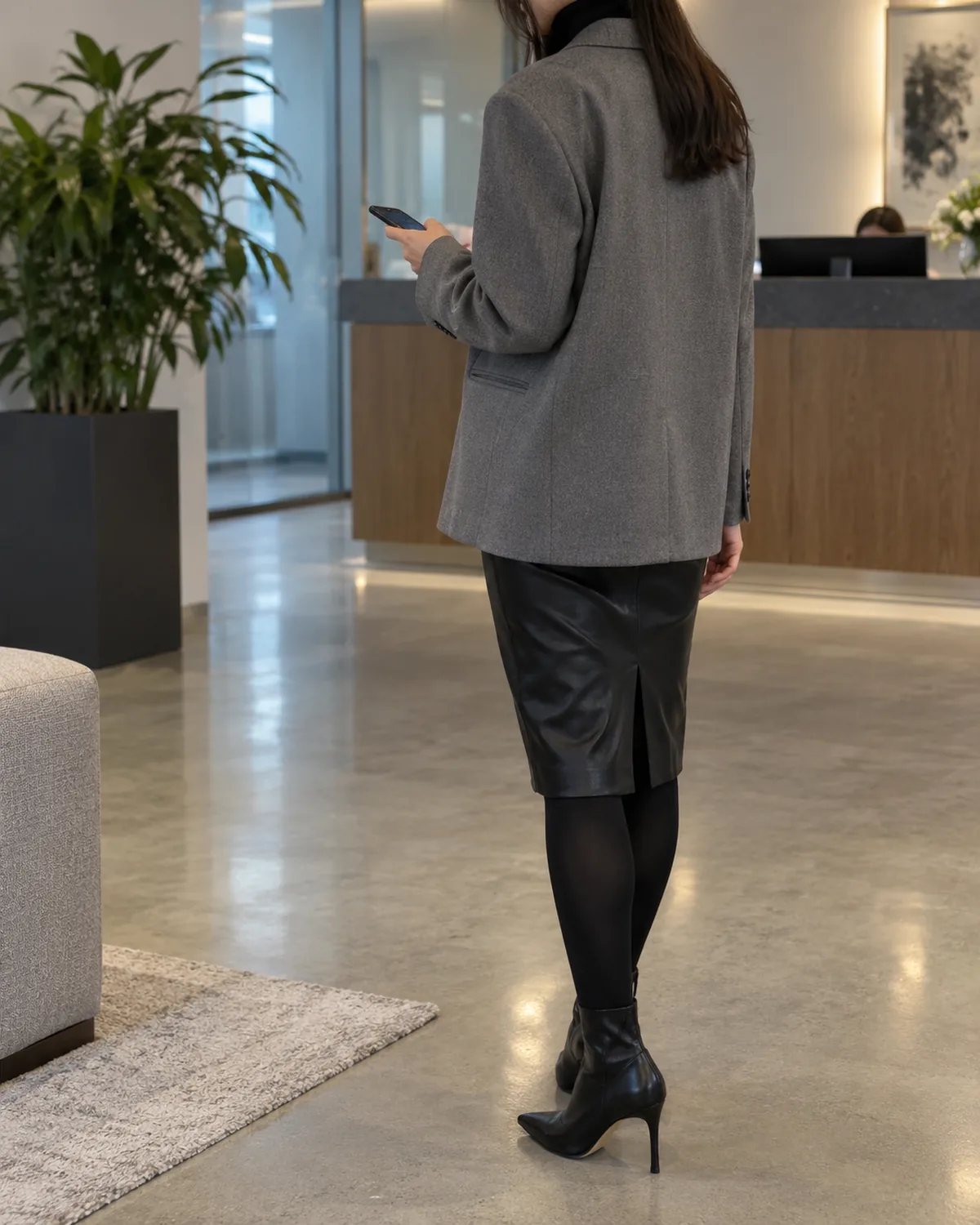 A woman at back-three-quarter angle looking down at a phone at waist level in an office reception area with a tall plant wearing a grey oversized blazer over a black fitted rollneck and black leather pencil skirt to the knee with black opaque tights and black pointed-toe ankle boots