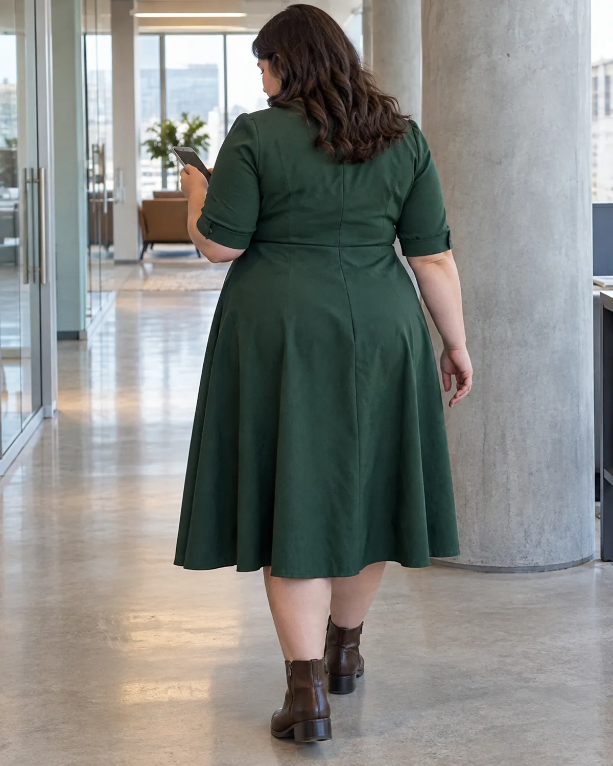 A plus-size woman seen from behind walking through an office corridor past a concrete column wearing a dark green button-front midi dress with elbow sleeves and an A-line skirt with brown leather ankle boots