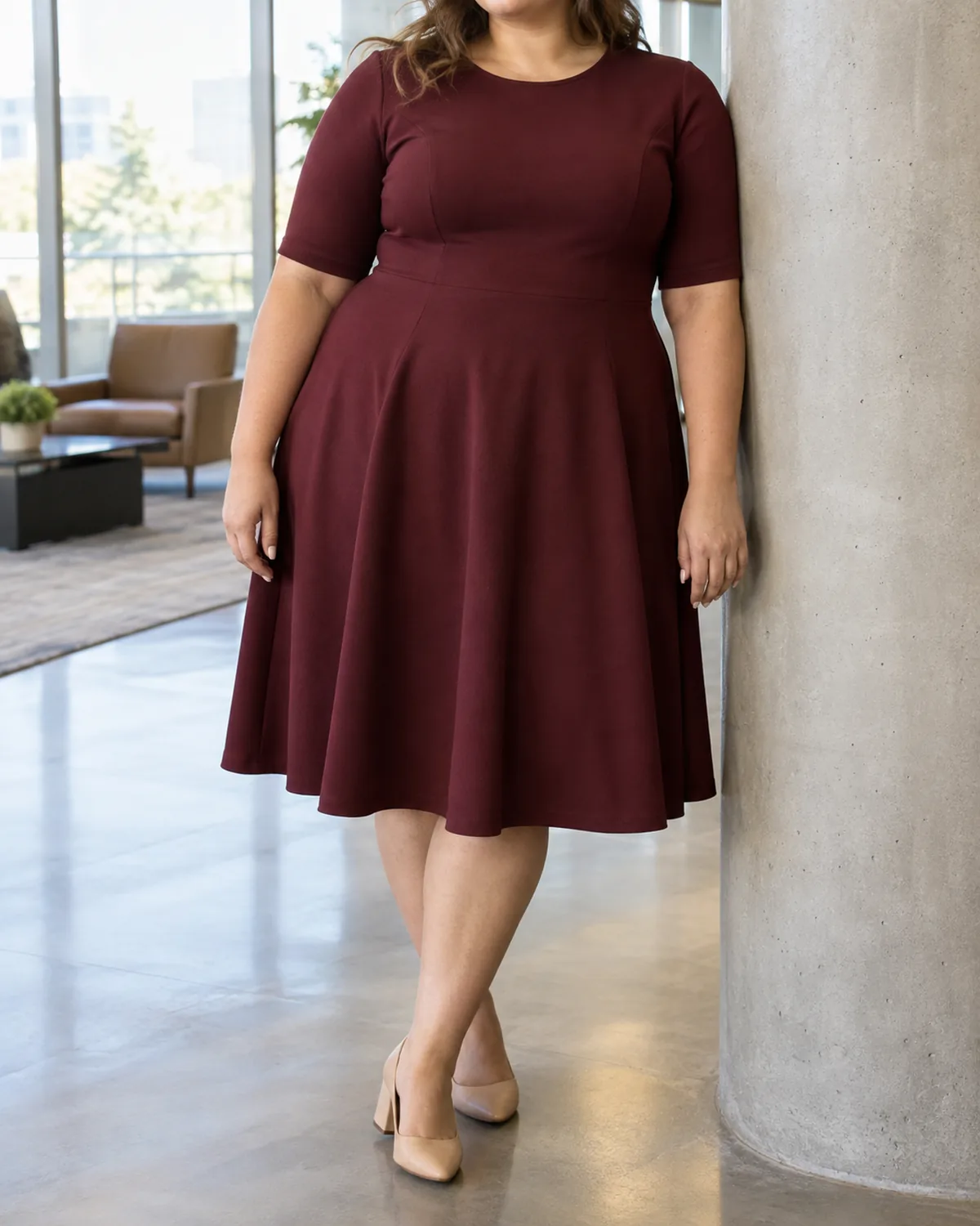 A plus-size woman leaning against a concrete column in an office lobby wearing a burgundy crew-neck fit-and-flare dress with elbow sleeves and a hem at the knee with nude pumps