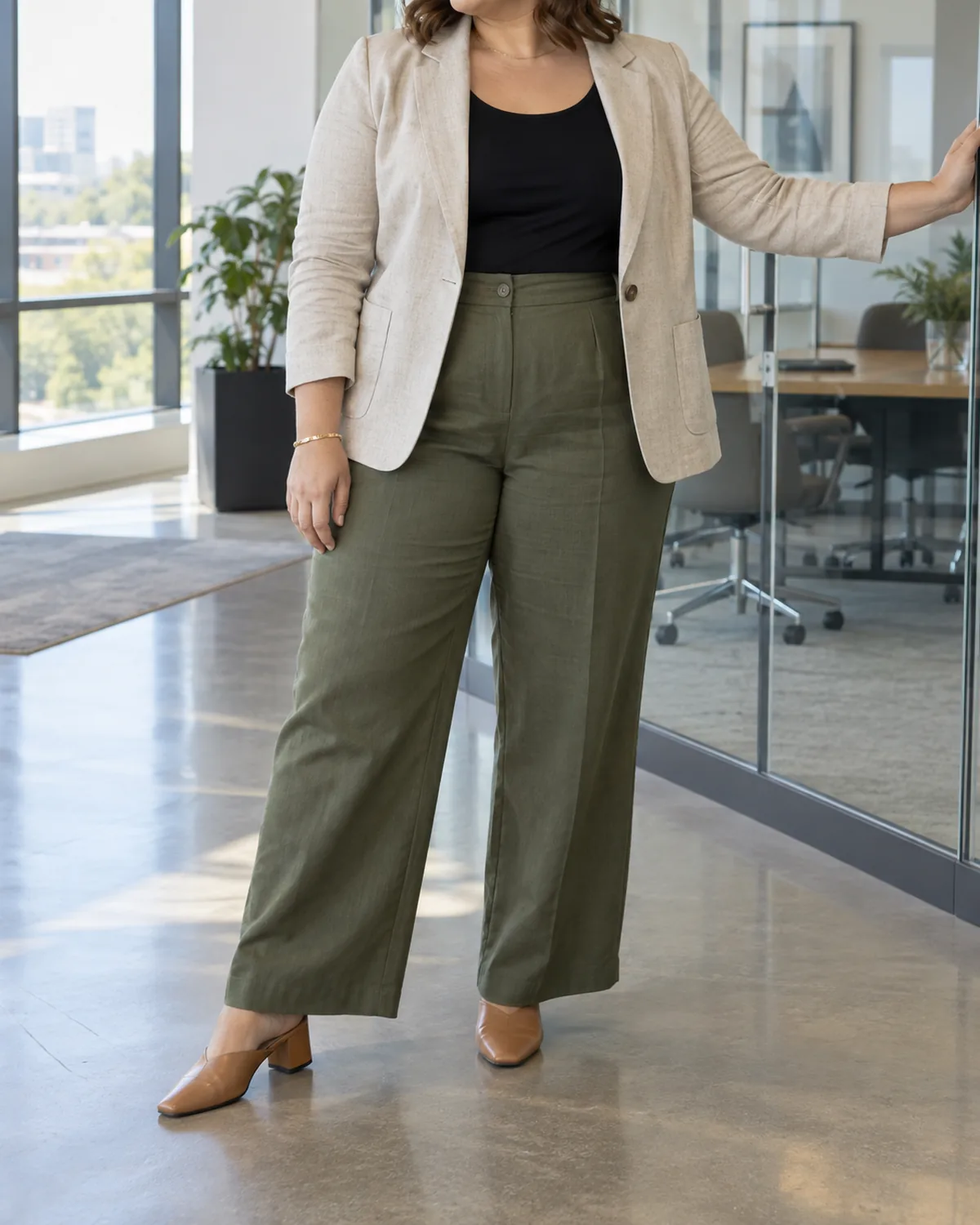 A plus-size woman standing in a modern office leaning one hand on a glass partition wearing an oatmeal linen-blend blazer over a black jersey shell with olive wide-leg trousers and tan block-heel mules