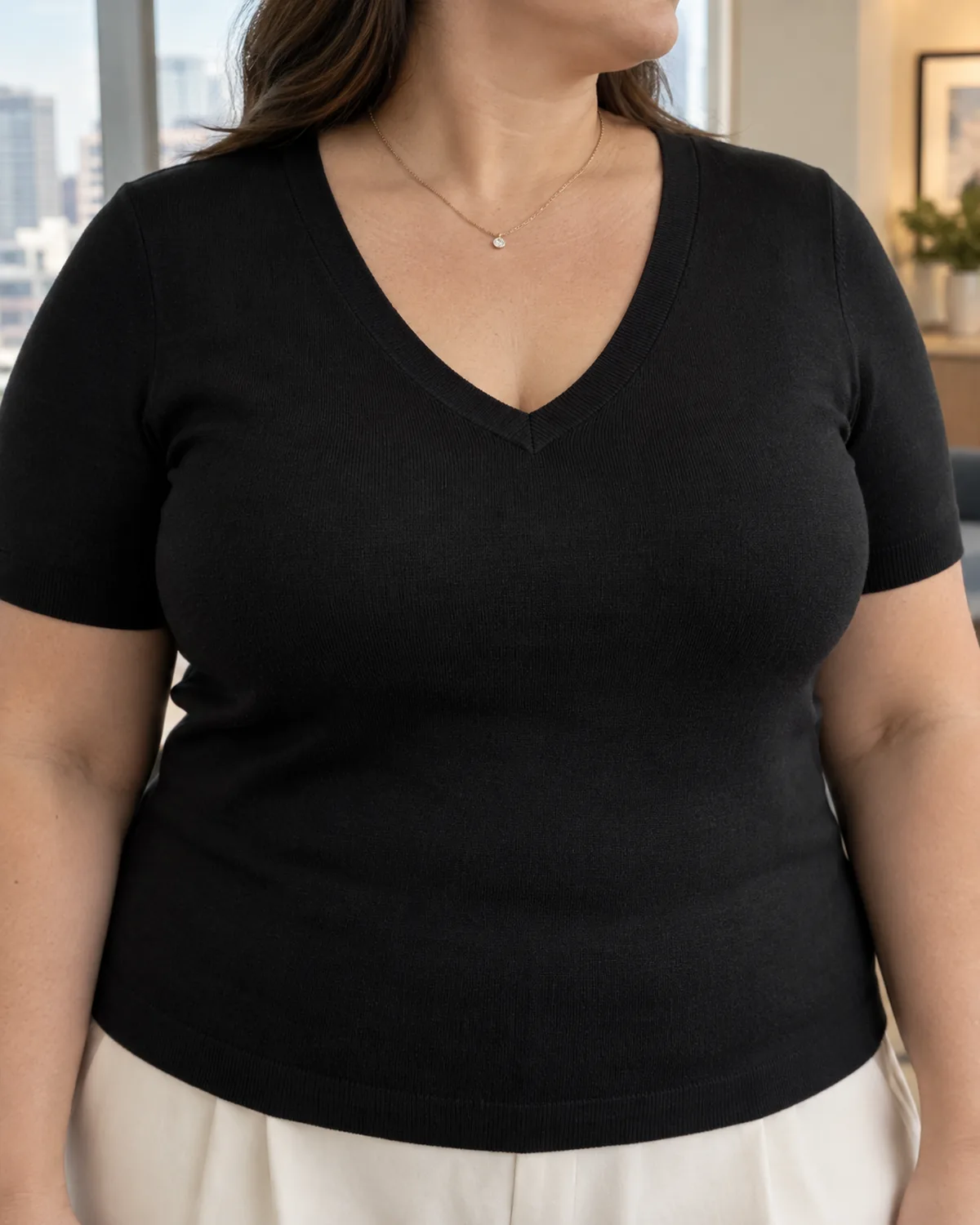Shoulder-to-waist close-up of a plus-size woman wearing a black fitted V-neck short-sleeve knit top with a delicate pendant necklace and the waistband of cream palazzo trousers visible at the lower edge