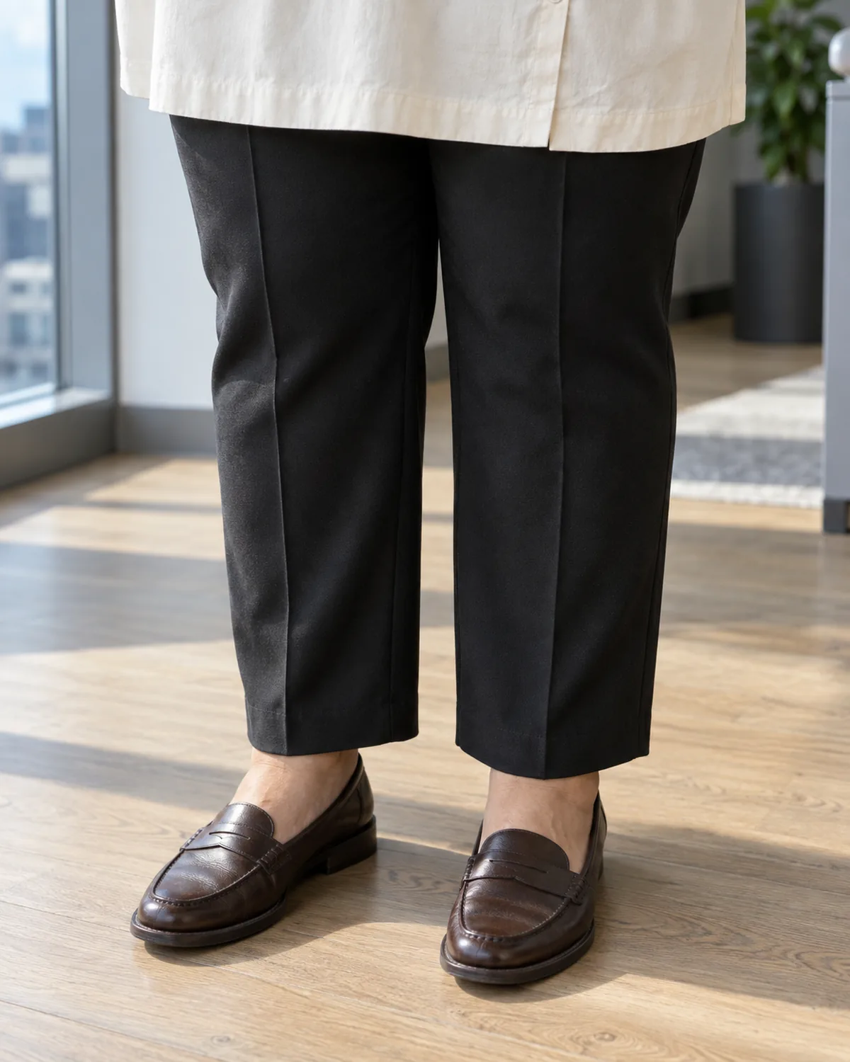 Knee-to-floor crop of a plus-size woman wearing black ponte trousers with a pressed centre crease and dark brown penny loafers with the hem of a cream tunic blouse visible at the top edge