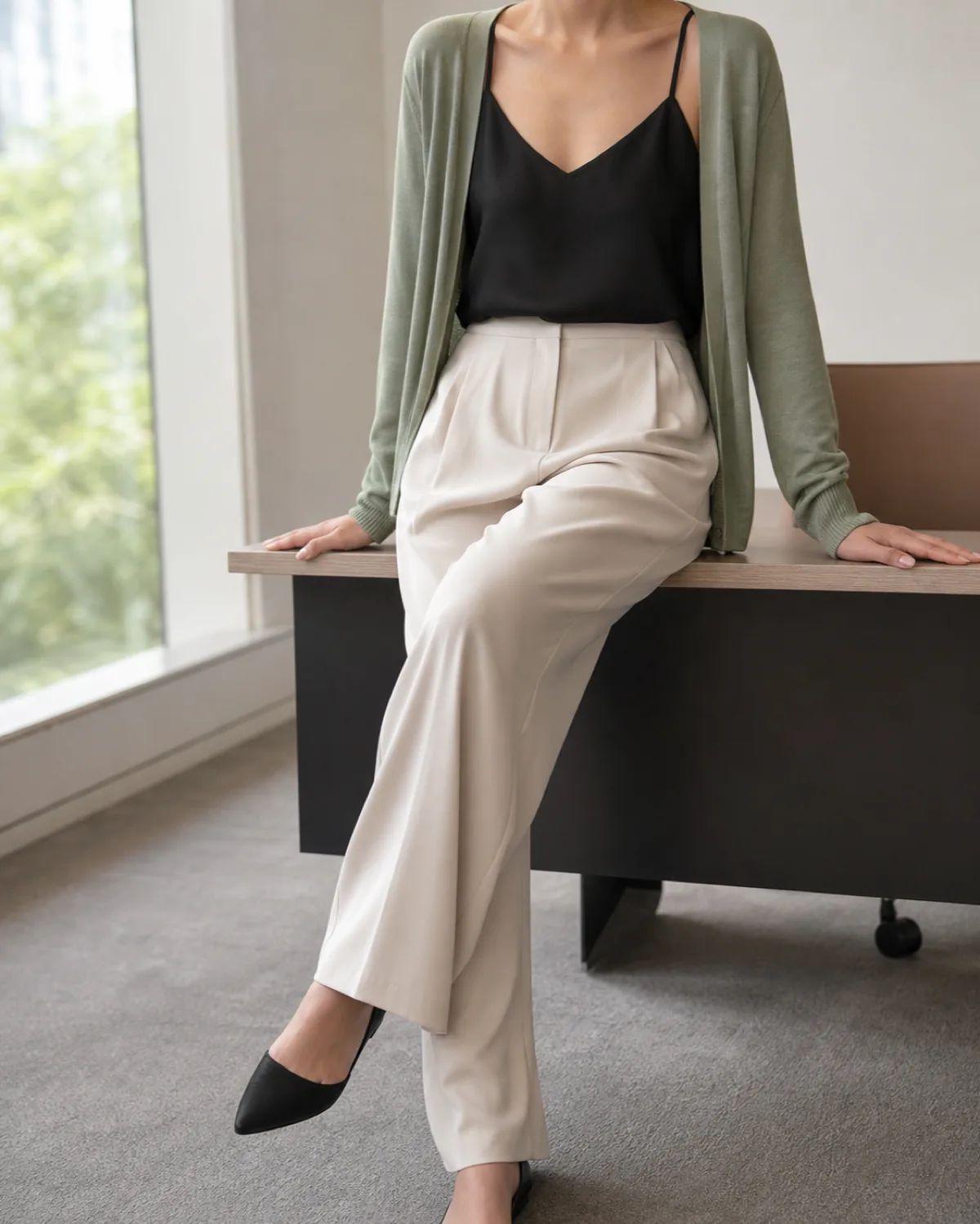 A woman seated on a desk edge in a sage green fine-gauge cardigan open over a black camisole with cream wide-leg trousers and black pointed-toe flats