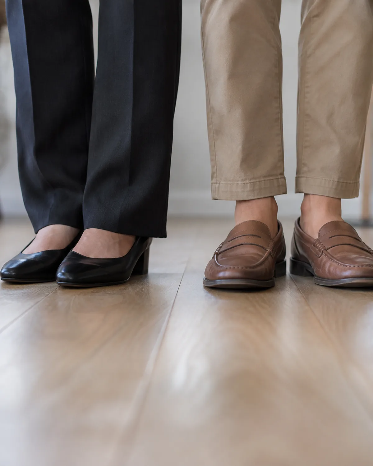 Low-angle close crop at floor level showing black closed-toe leather pumps with dark trouser hems on the left and tan leather loafers with khaki chino hems showing the ankle on the right