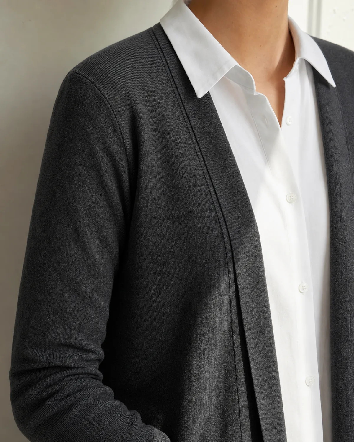 Close crop on the shoulder and collar line of a longline charcoal fine knit cardigan worn open over a white collared shirt