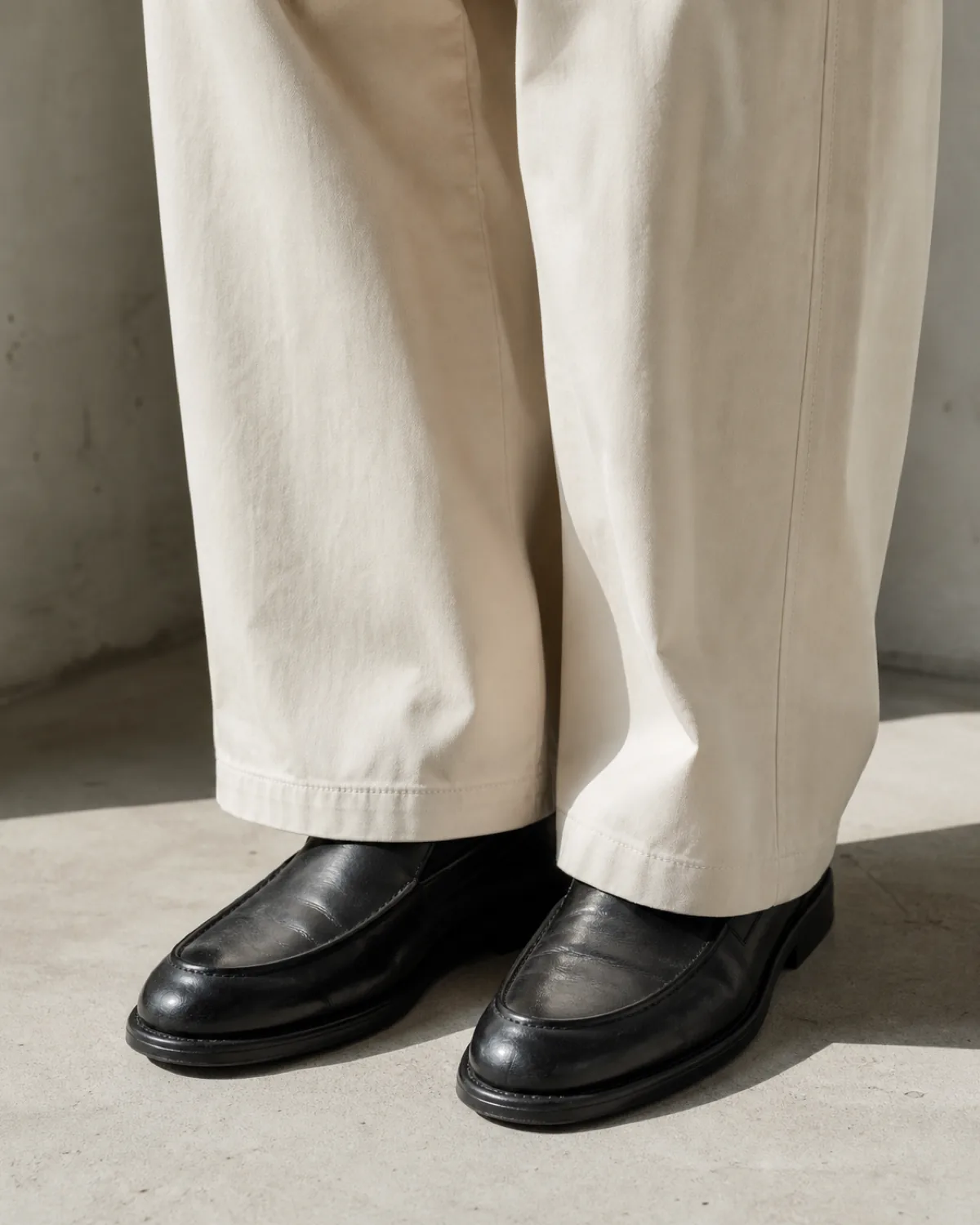 Cream wide-leg trousers breaking just above plain black leather loafers on a pale concrete floor