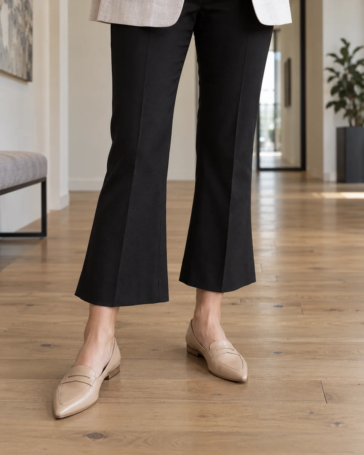 Close crop from knee to floor showing black cropped kick-flare trousers ending just above the ankle with nude pointed-toe leather loafers and the hem of a linen blazer visible at the top of the frame on a light hardwood office floor