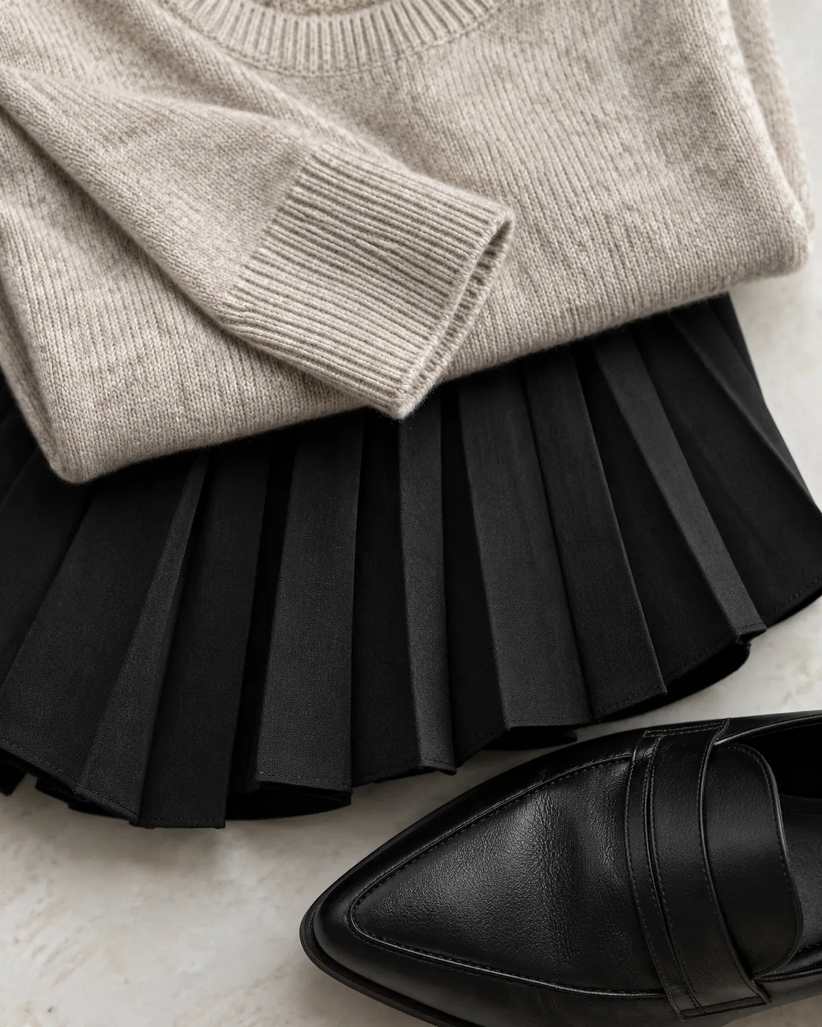 Stacked detail macro of three overlapping elements: an oatmeal crewneck knit sweater with ribbed cuffs, a section of black knife-pleat midi skirt fabric showing sharp pleat edges, and the toe of a black pointed leather loafer