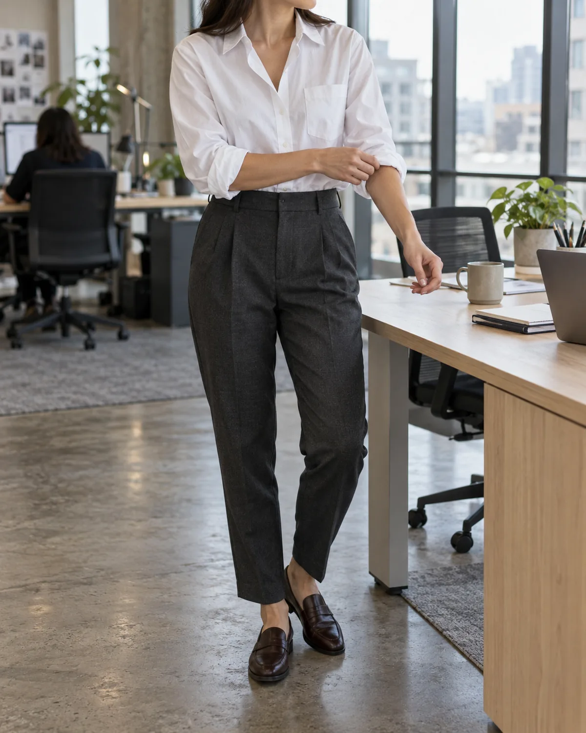 A woman standing beside a light wood desk in an open-plan office, mid-torso crop from chin to knee, adjusting the cuff of an oversized white cotton poplin shirt front-tucked into charcoal tapered trousers with dark brown leather loafers