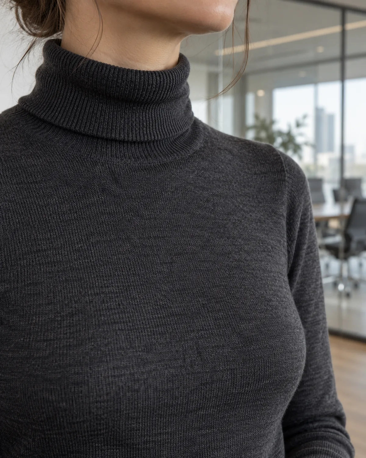 Close crop on the shoulder and collar area of a woman wearing a charcoal fine-gauge turtleneck with visible ribbed knit texture at the folded collar, with a softly blurred glass-walled conference room behind