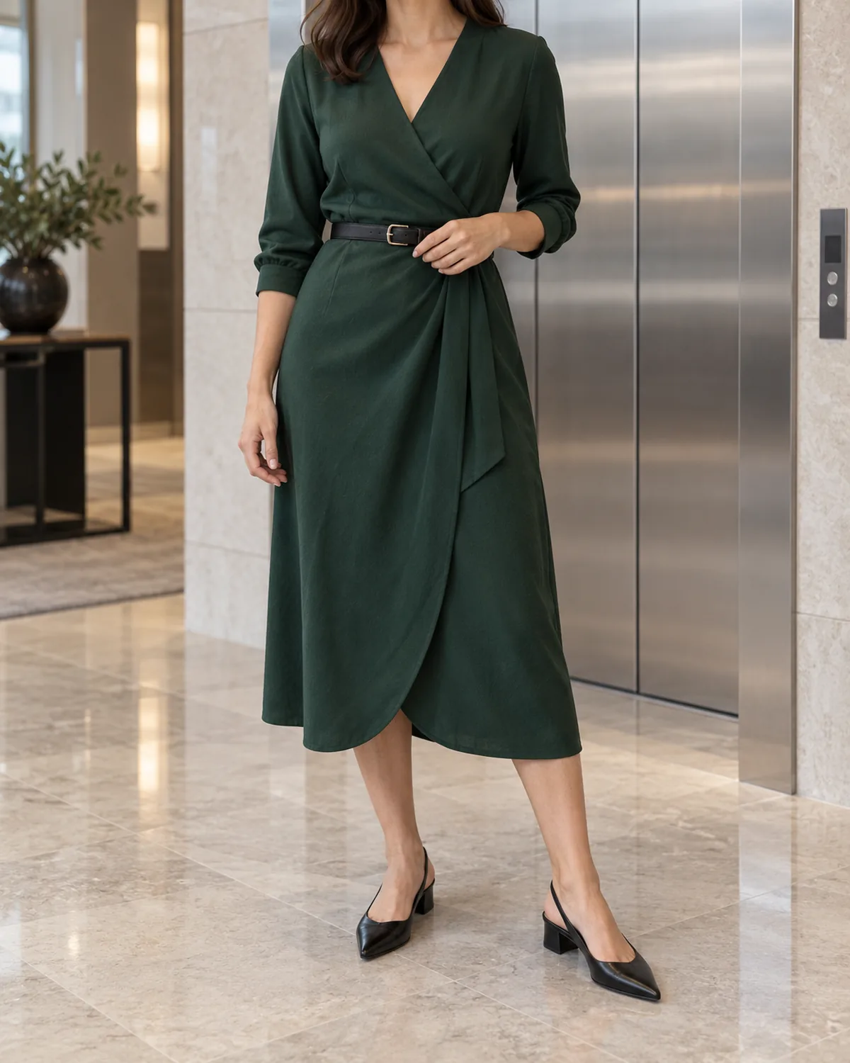A woman standing in an elevator lobby with stainless steel doors behind her wearing a forest-green midi wrap dress cinched at the waist with a thin black leather belt and black low-heel slingback shoes