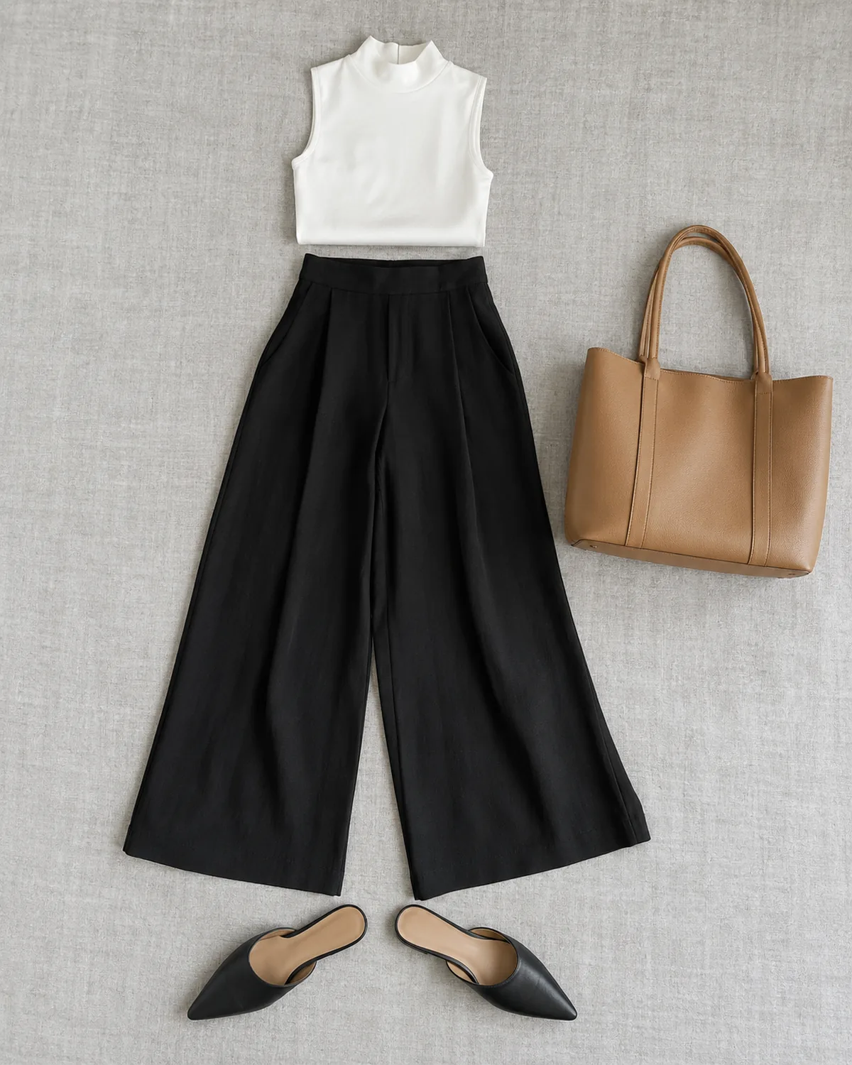 Overhead flat-lay of a white mock-neck sleeveless shell and black wide-leg crepe trousers on a grey linen surface with a tan leather tote and black pointed-toe mules