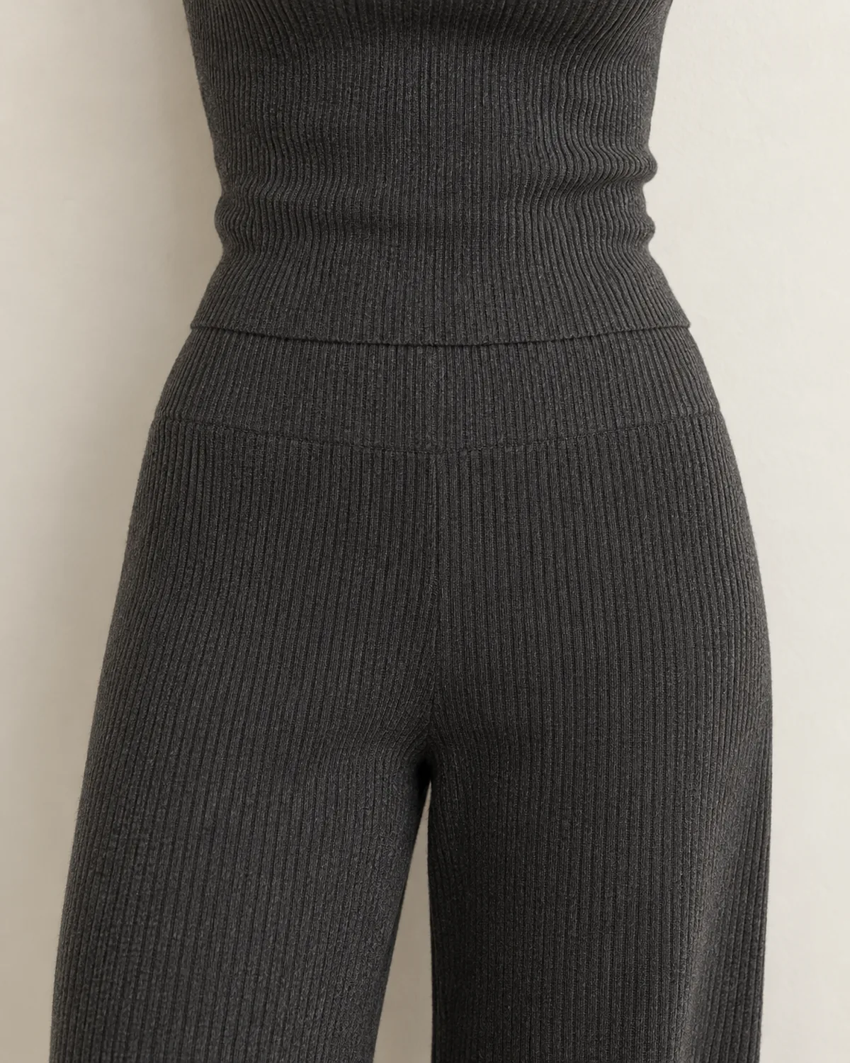 Close crop on the waist of a woman wearing a matching charcoal ribbed knit trouser set showing the seamless transition where the fitted crew-neck knit top meets the high-waist pull-on trousers