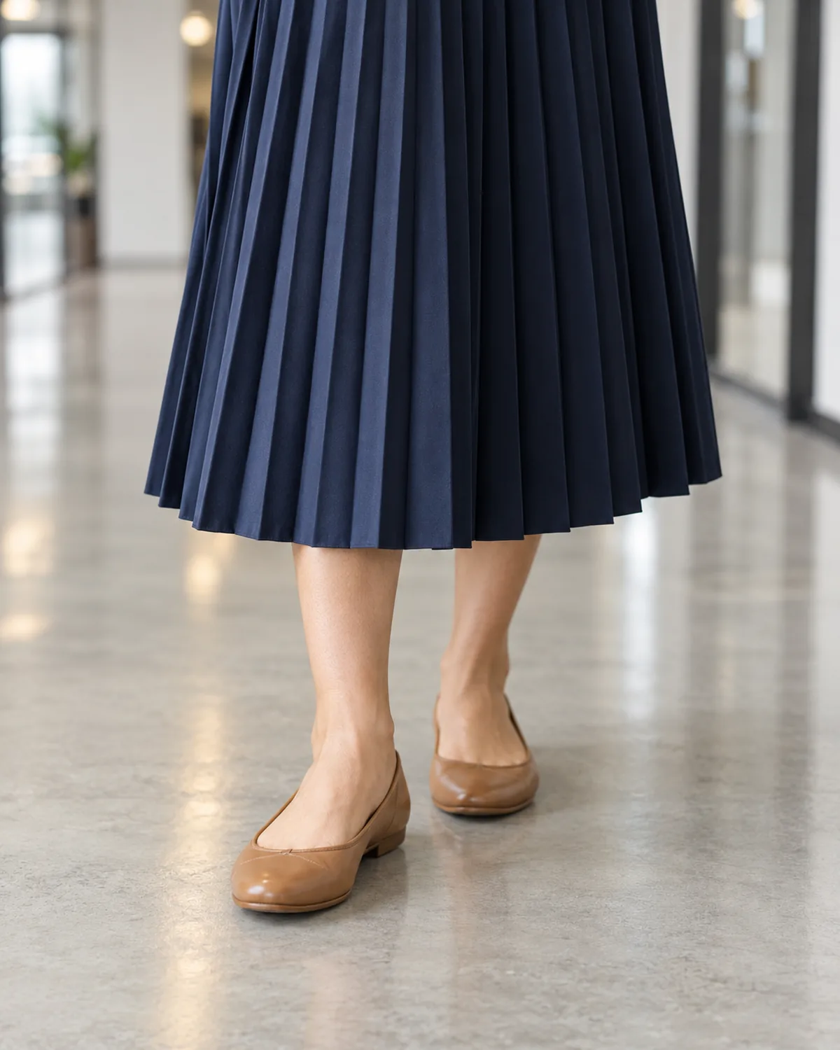 Close crop from the knee down showing a navy knife-pleated midi skirt hem and tan cushioned leather ballet flats on a polished grey concrete floor