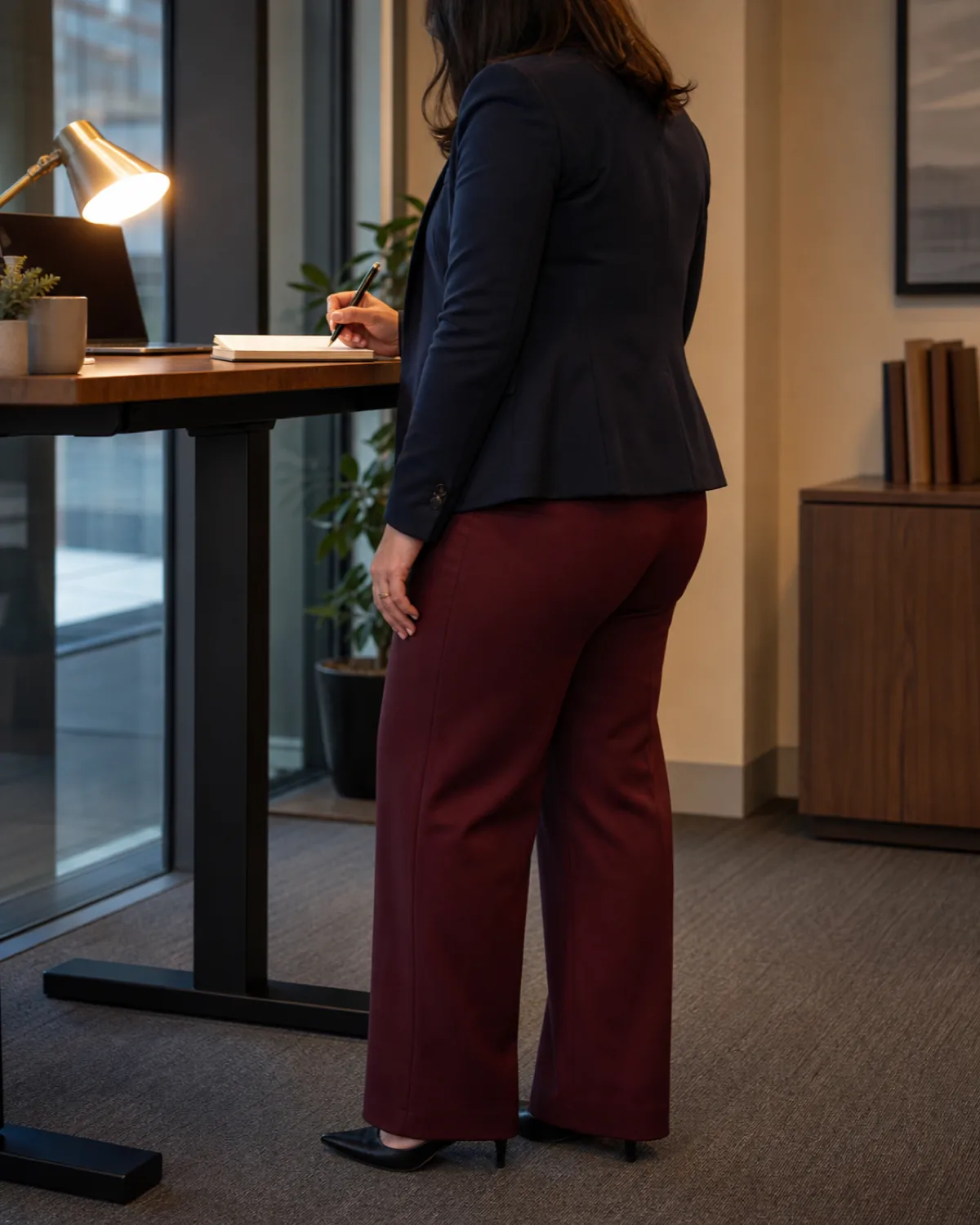 A midsize woman at a standing desk from a back three-quarter angle wearing burgundy wide-leg trousers, a navy blazer, and writing in a notebook