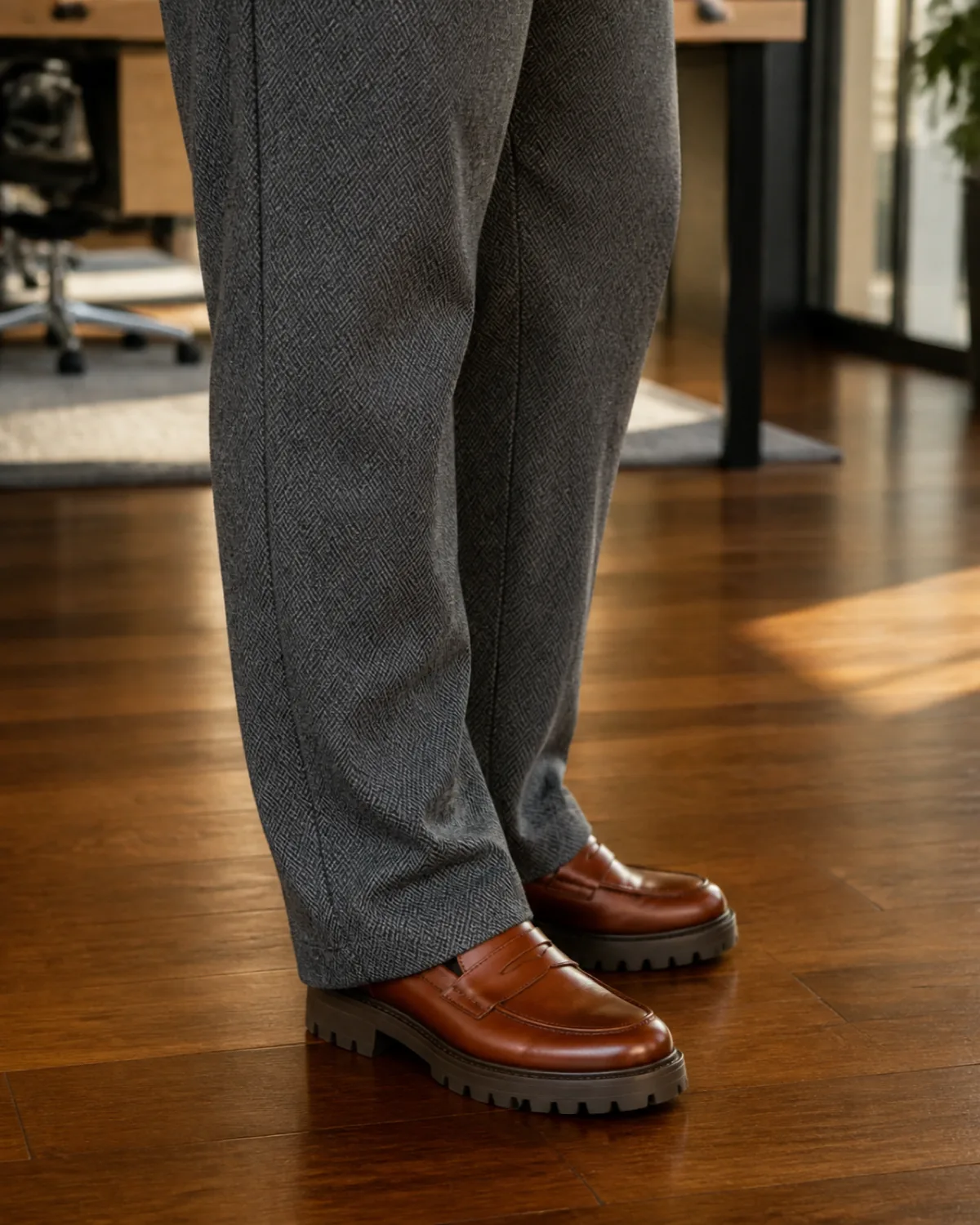 Close crop on grey herringbone wool trousers breaking cleanly at cognac chunky loafers on a hardwood office floor