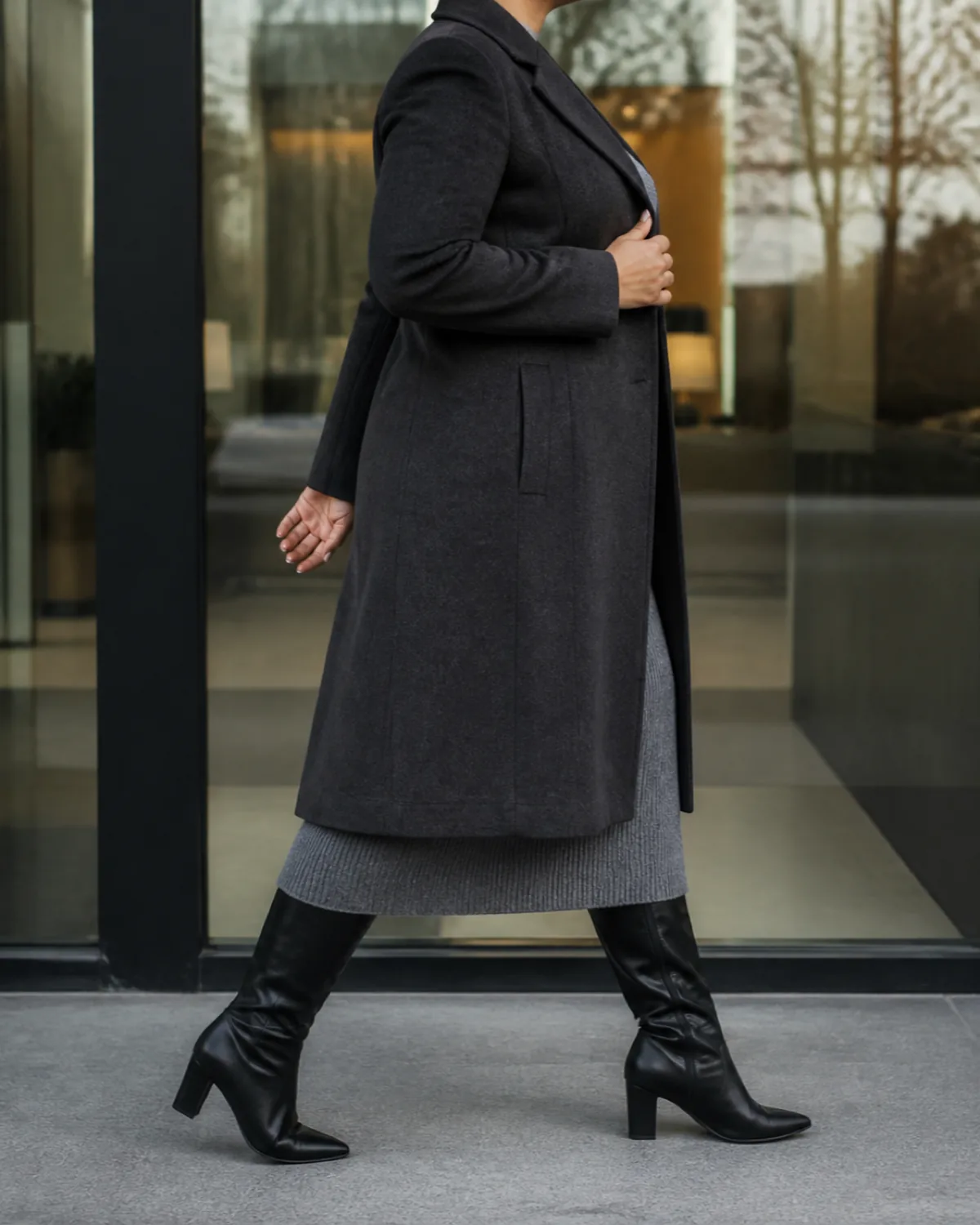 A midsize woman walking in profile wearing a charcoal longline wool coat over a grey knit midi dress with black knee-high boots