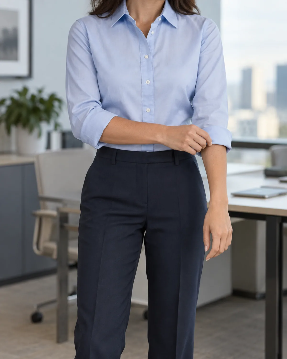 Mid-torso crop of a woman adjusting her right shirt cuff with her left hand wearing a light blue button-down shirt with sleeves rolled once tucked into dark navy trousers with a sharp front crease in an office setting