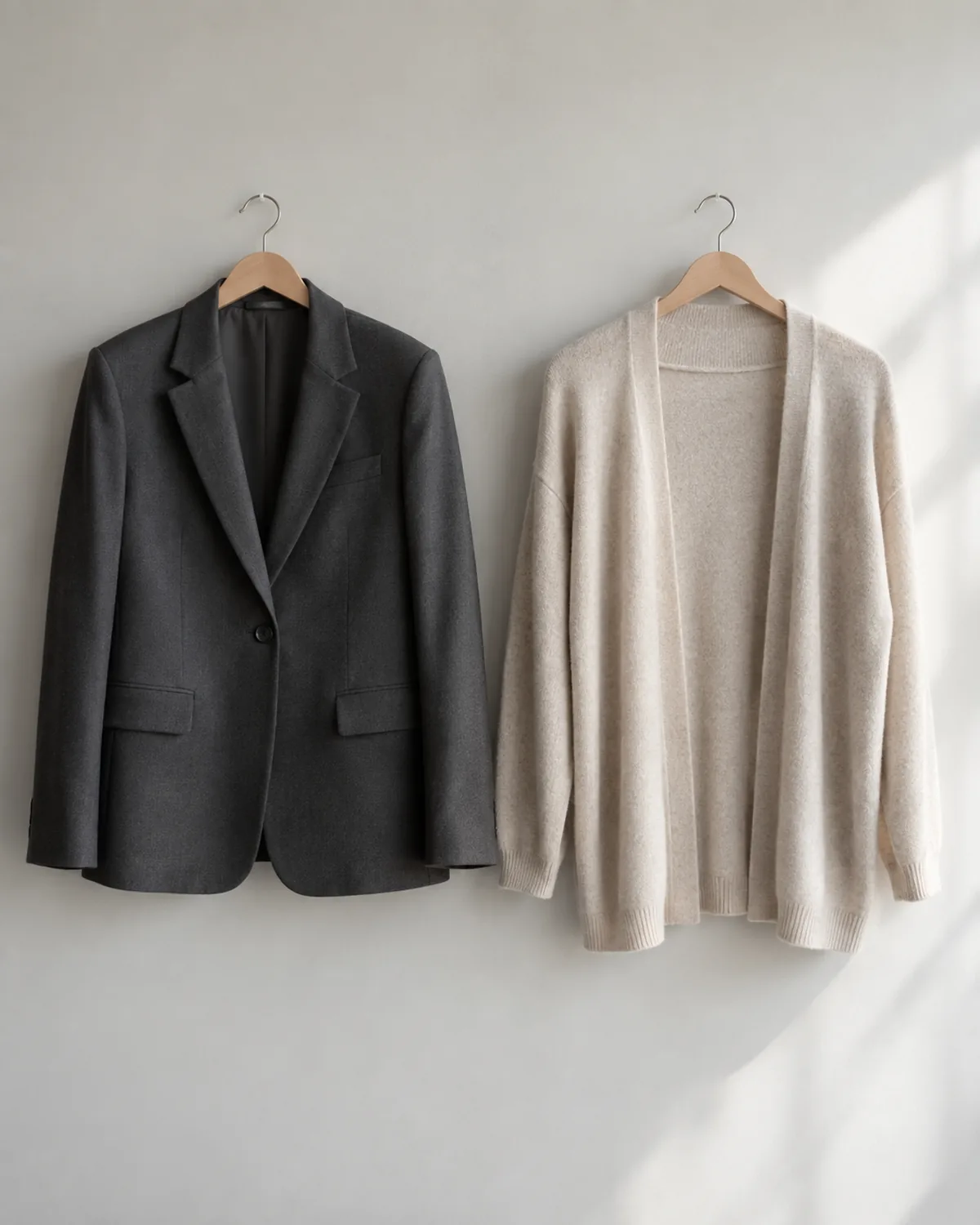 A dark charcoal structured single-breasted blazer and a soft oatmeal fine-gauge knit open-front cardigan hanging side by side on wooden hangers against a light grey wall showing two valid work layers at different formality levels
