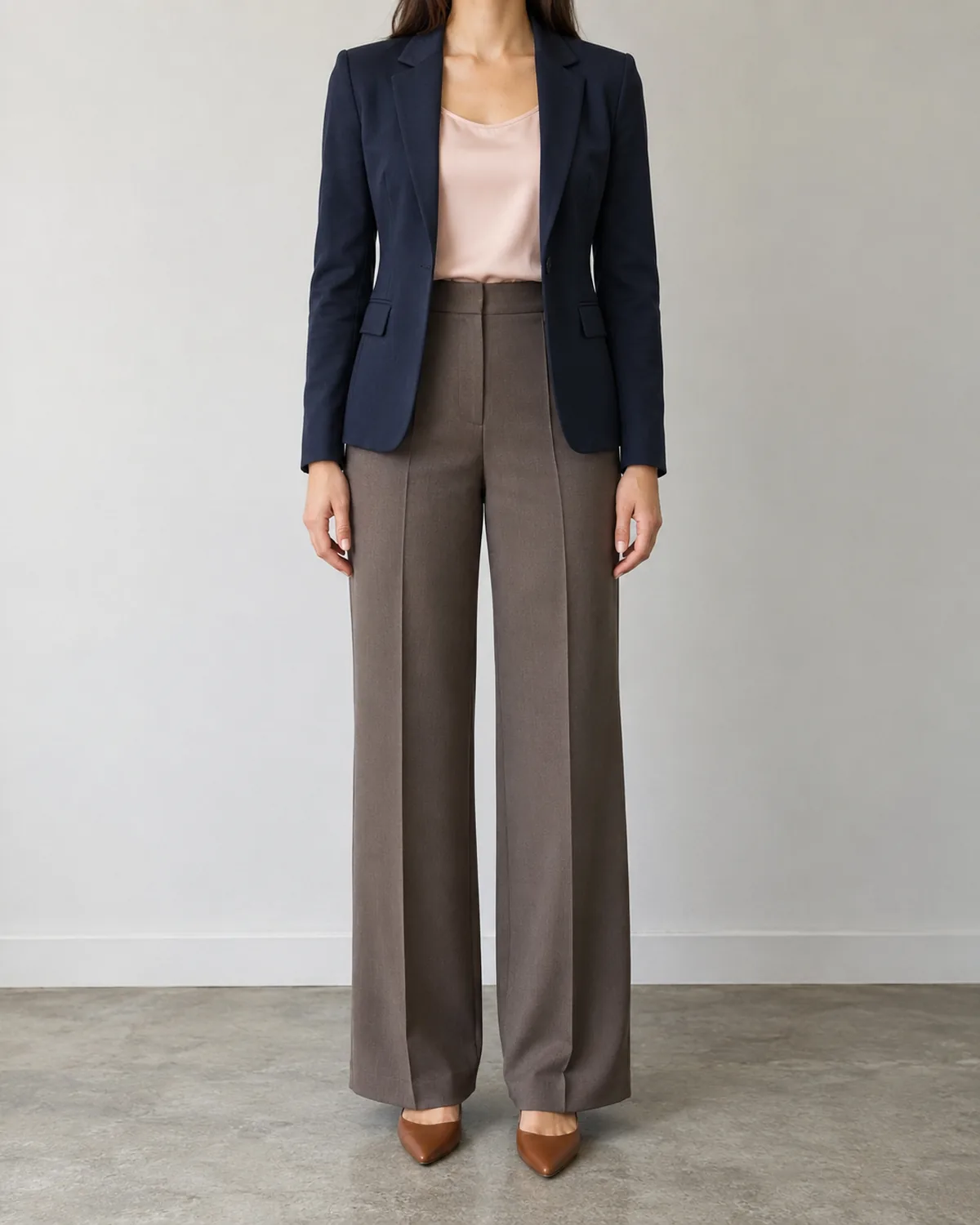 A woman standing straight-on in front of a grey office wall wearing a fitted navy ponte blazer over a soft blush silk camisole tucked into dark taupe wide-leg trousers with cognac pointed-toe flats