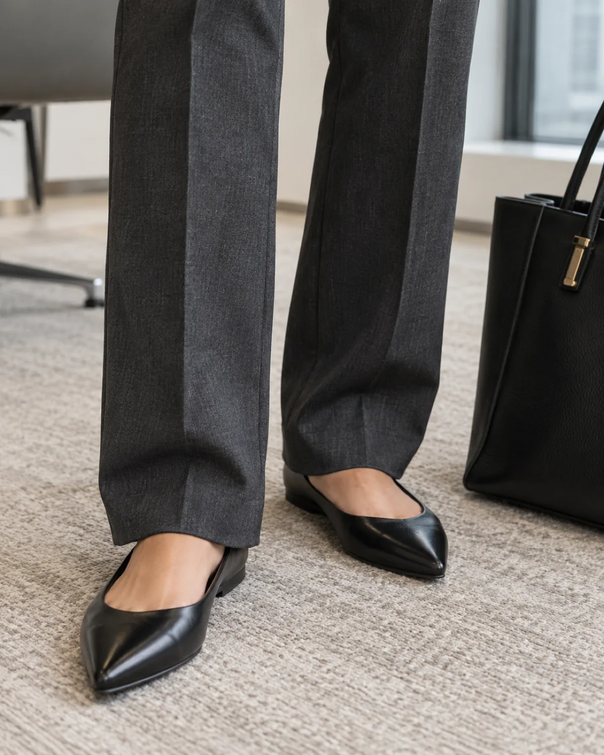 Close crop from mid-calf to the floor of dark charcoal straight-leg trousers breaking slightly over black pointed-toe leather flats with a structured black leather tote bag with gold-tone hardware on the floor beside the right foot