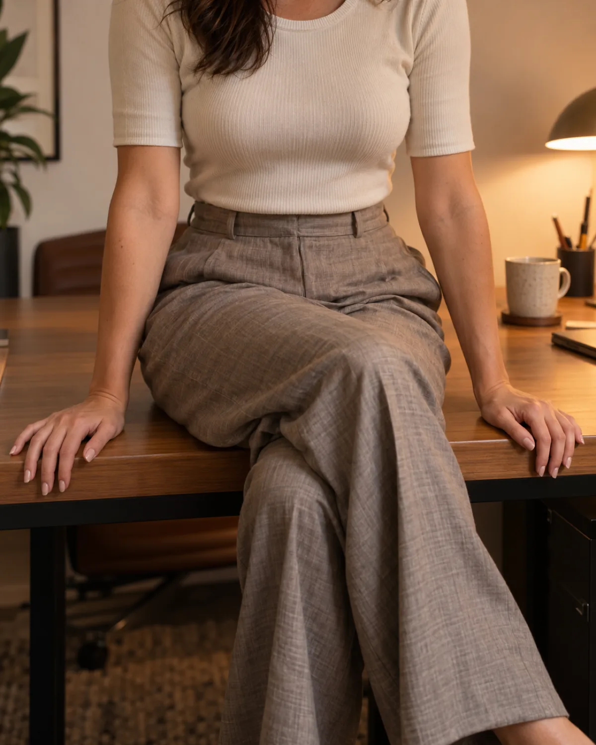 A woman seated on the edge of an office desk with legs crossed wearing taupe wide-leg linen trousers that are bunching and pooling at the knee and around the desk edge with a cream fitted knit top tucked in