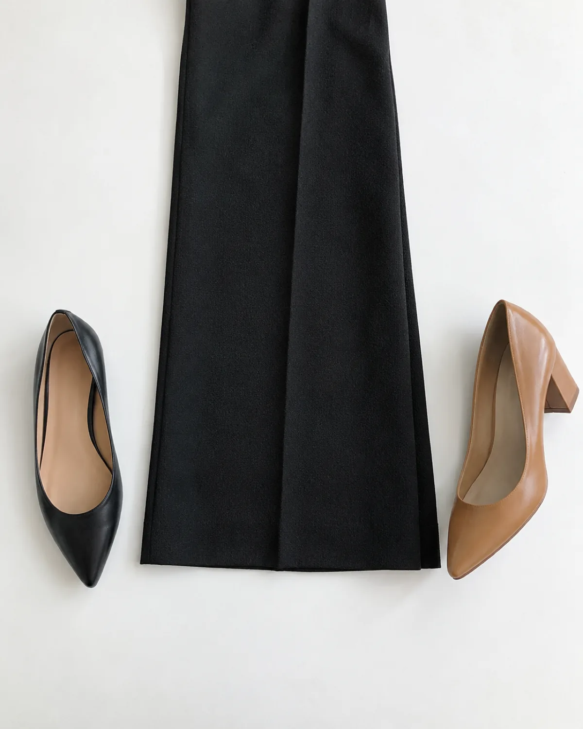 Overhead flat-lay on a white surface showing a black wide-leg wool trouser leg laid flat with a black leather pointed-toe ballet flat to the left and a tan leather block-heel pump to the right