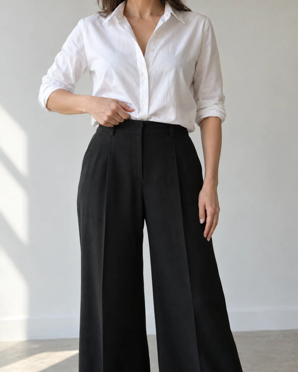 A woman from chin to below the knee standing straight-on with her right hand pushing the center-front of a white cotton poplin shirt into the waistband of black wide-leg wool trousers in a front-tuck in progress with her left hand at her side