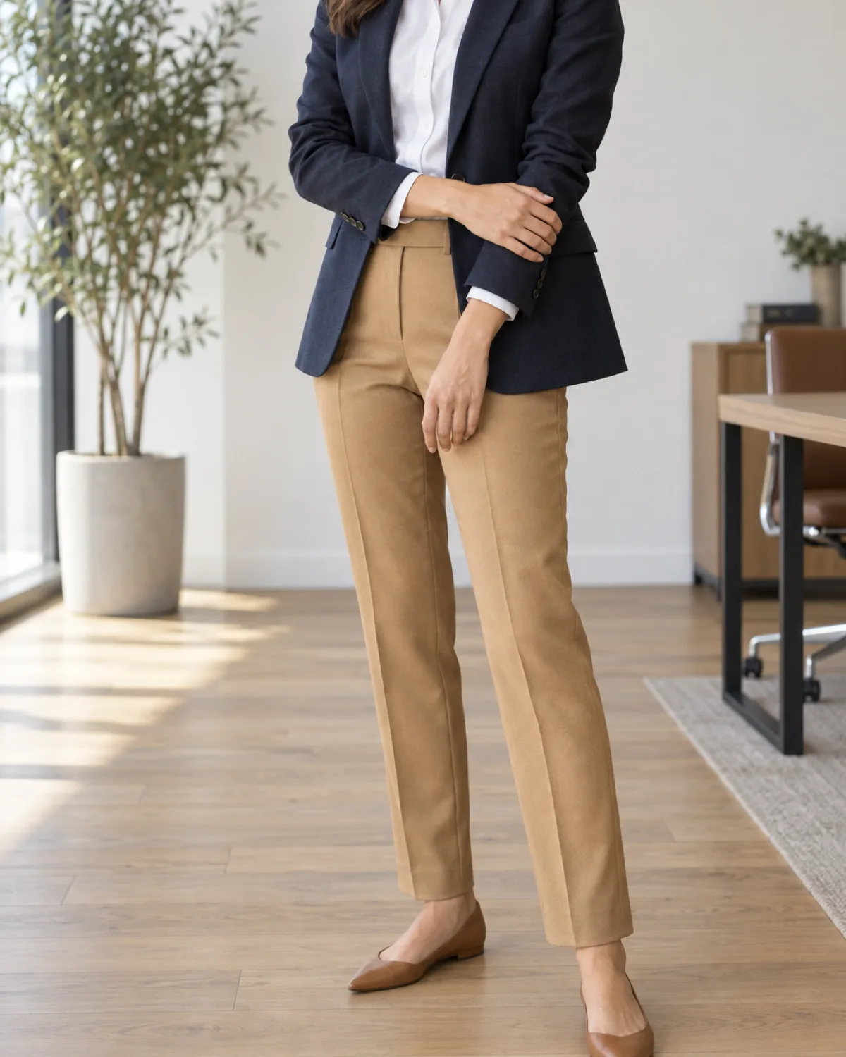 A woman standing in a three-quarter turn wearing warm camel tailored trousers with a front crease, a white cotton shirt tucked in, a navy single-breasted blazer, and tan pointed-toe flats, in a modern office with pale oak flooring and indoor plant