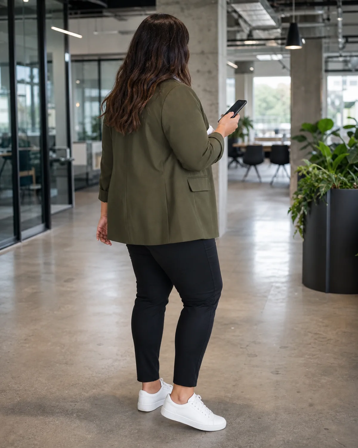 A plus-size woman viewed from behind in an olive column blazer with unstructured shoulders over black slim ankle pants and white sneakers, looking at a phone in a modern tech office with concrete and greenery