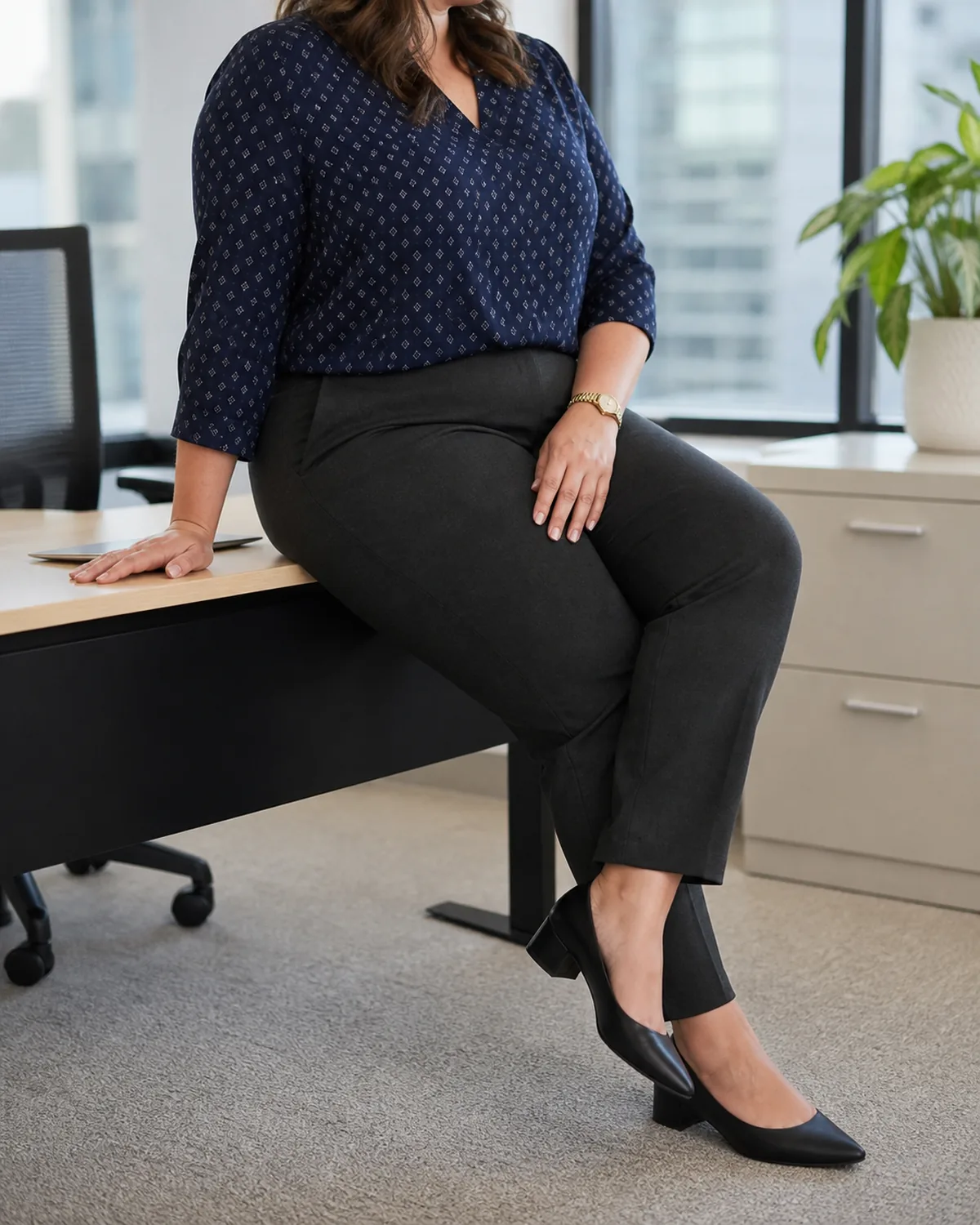 A plus-size woman seated at a desk edge in dark charcoal slim trousers and a navy V-neck blouse with a subtle geometric print, a gold watch on her wrist, one hand resting on the desk surface