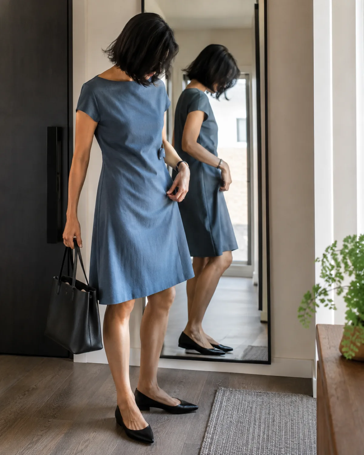 A woman in a slate blue A-line cotton dress standing near a mirror in a home entryway