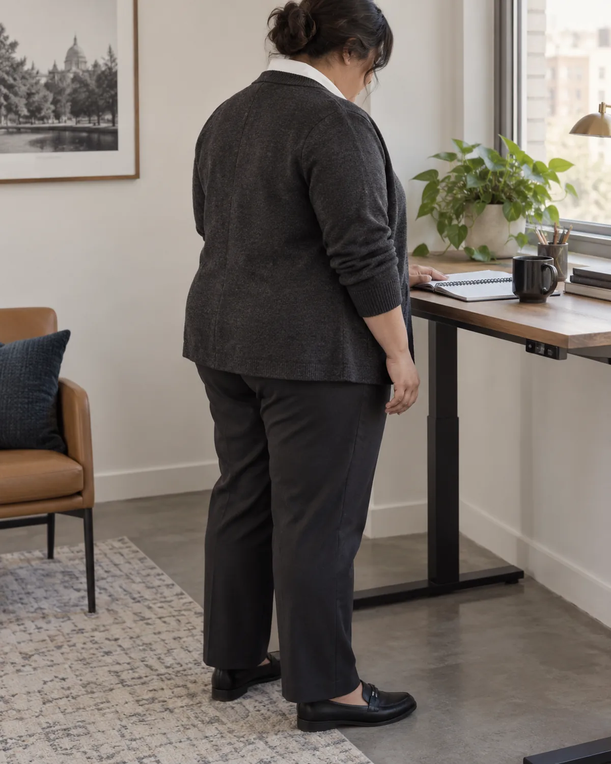 A plus size woman seen from the back-three-quarter angle looking down at a notebook on a desk, wearing a charcoal structured cardigan over a collared shirt and dark trousers