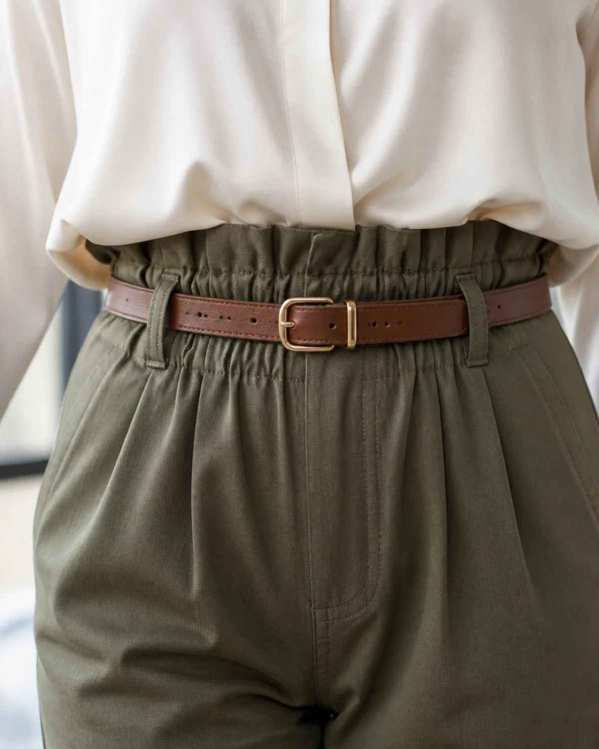 A woman in olive paperbag-waist trousers with a gathered waistband and a white blouse French-tucked at centre front, a thin tan leather belt visible, standing against a white brick wall