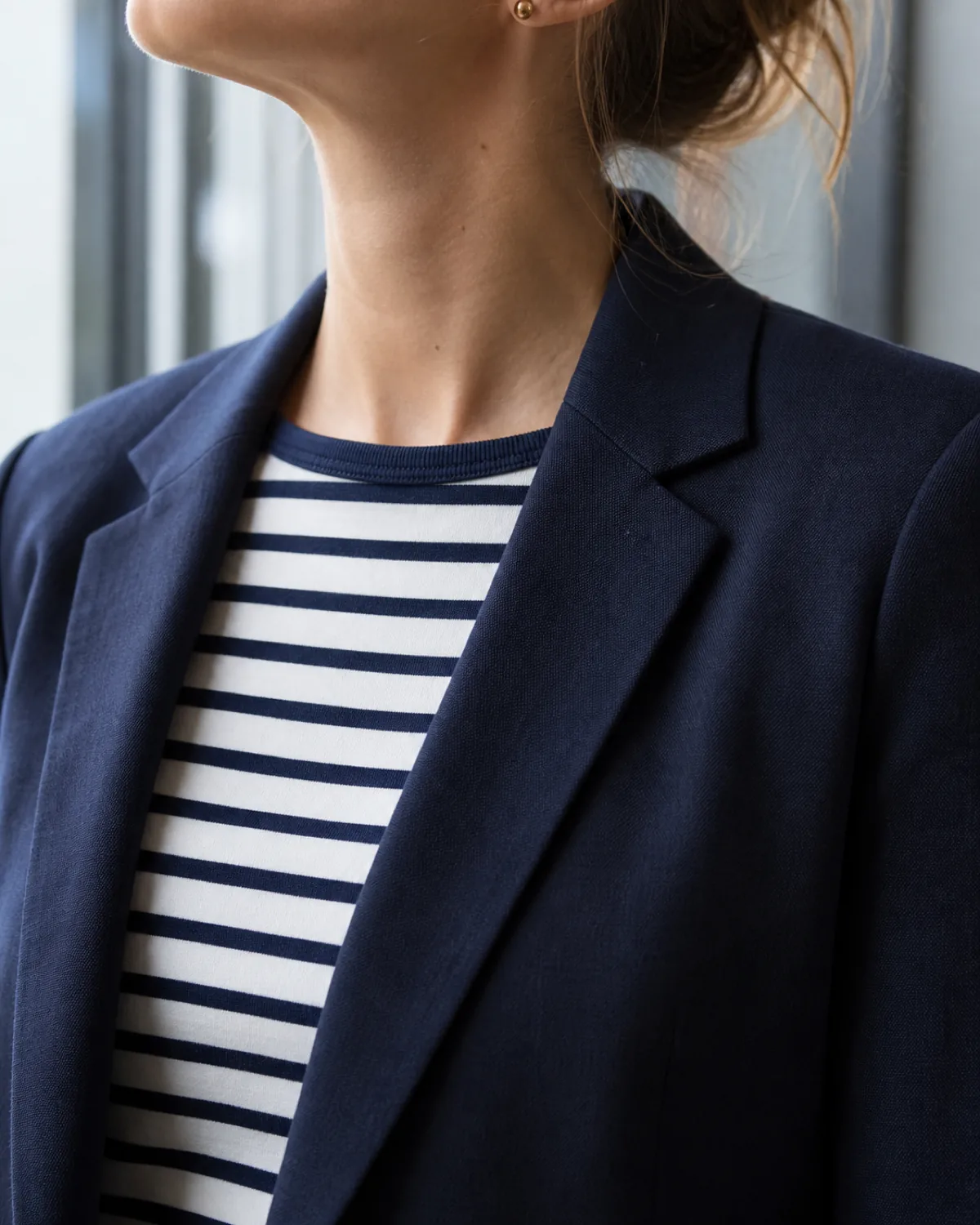 Close-up of a navy and white Breton-stripe tee under a navy blazer with notch lapels, cropped at the shoulder and collar with a small gold stud earring visible