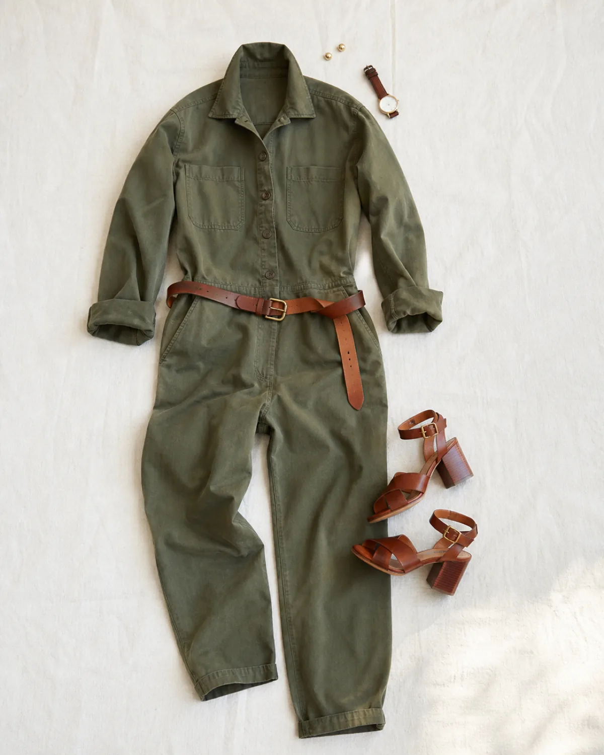 A flat-lay of an olive utility jumpsuit with a collar and patch pockets, a cognac leather belt, brown block-heel sandals, small gold stud earrings, and a watch on a white surface