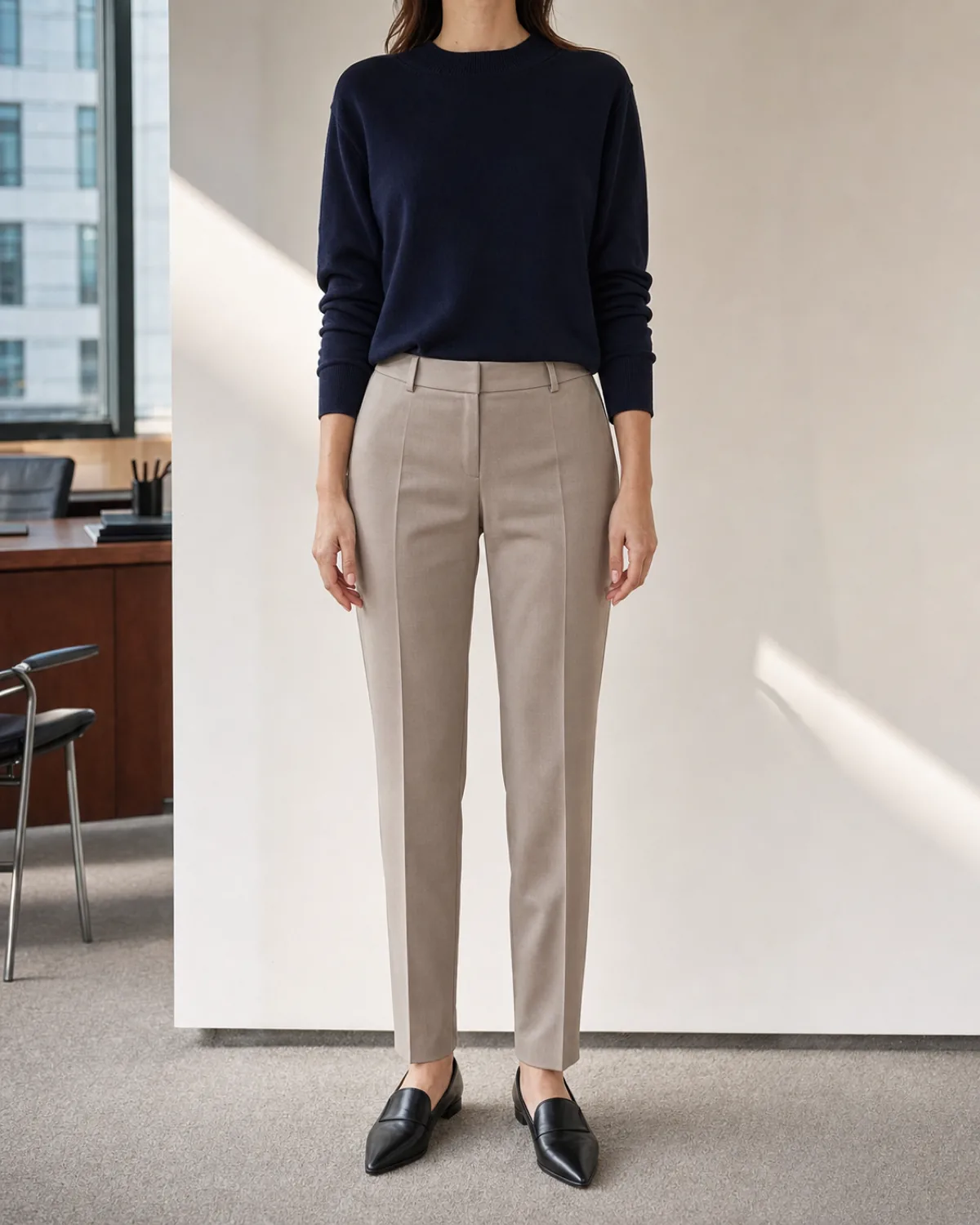 A woman standing straight-on in a navy crewneck knit tucked into stone tailored trousers with black pointed-toe loafers in a modern office