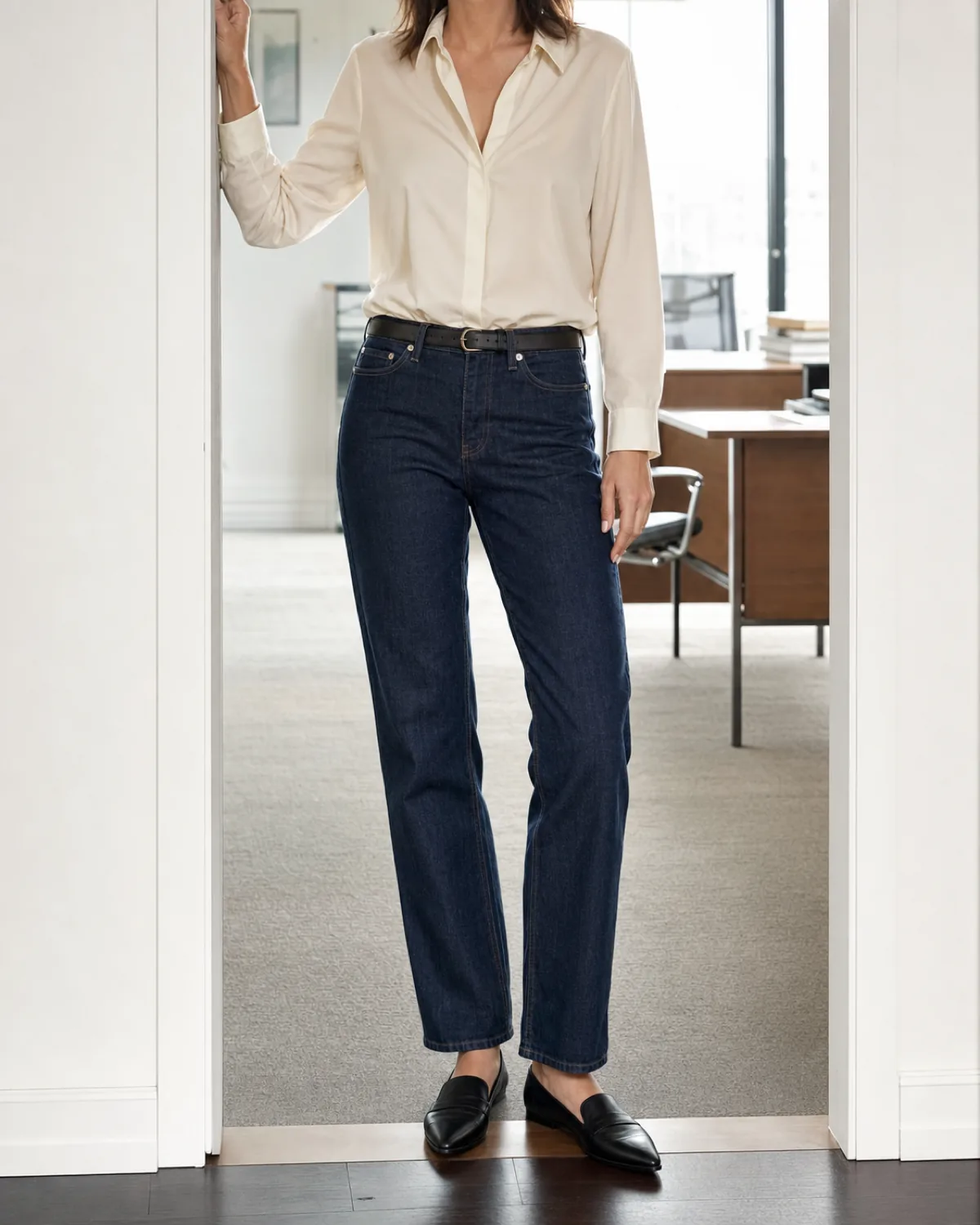 A woman standing in an office doorway in dark indigo straight-leg jeans with a cream silk blouse tucked in at the waist, a dark leather belt and black leather loafers