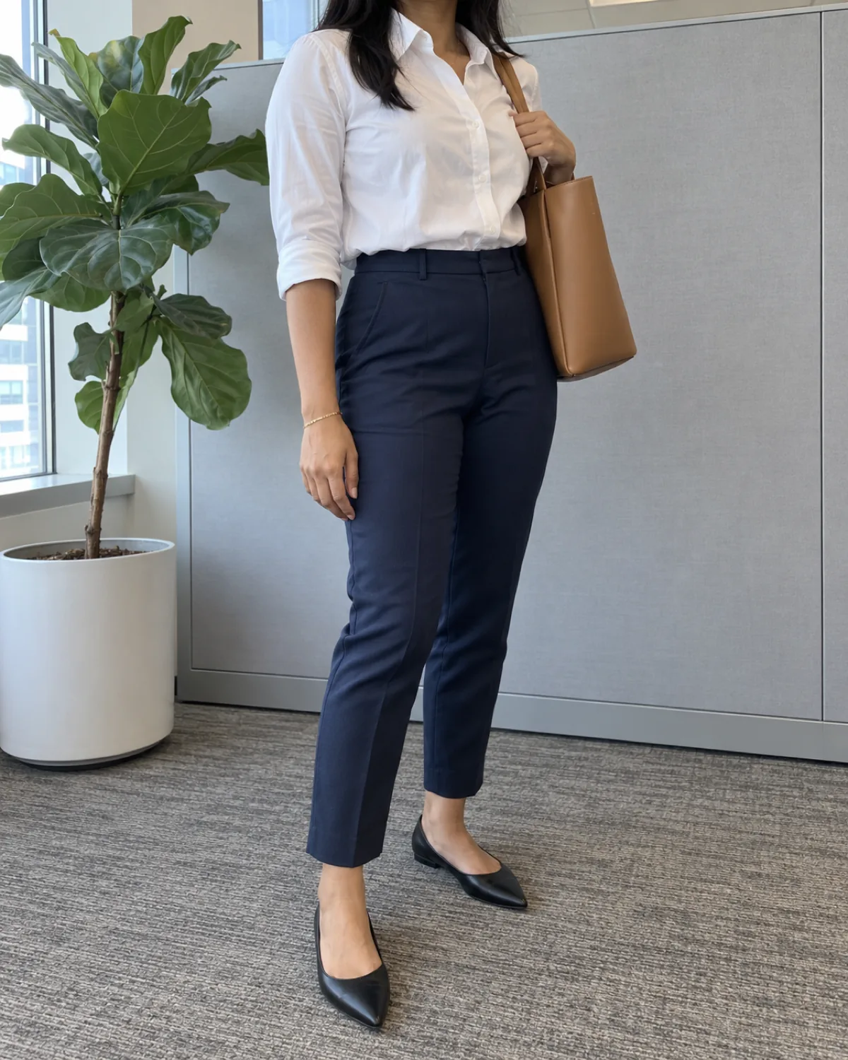 A woman in a three-quarter turn wearing navy high-waisted ankle trousers with a tucked white button-up blouse and a tan structured tote on her shoulder with black pointed-toe flats in front of a grey office partition and a fiddle leaf fig