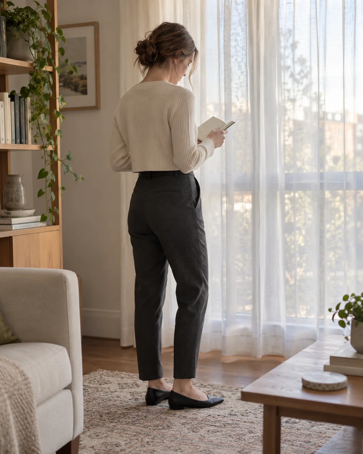 A woman seen from a back-three-quarter angle reading a small notebook wearing a cream ribbed cropped cardigan ending at the waistband of charcoal tapered trousers with black flats