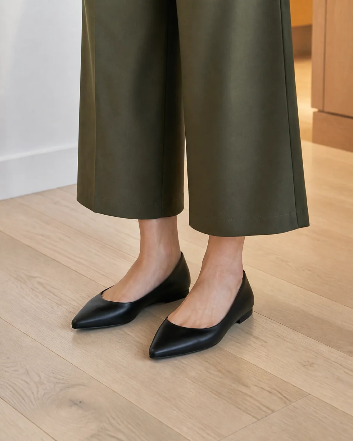 Close crop on the lower legs of a petite woman standing on light hardwood floor showing olive cropped wide-leg trouser hems above bare ankles and black pointed-toe flats