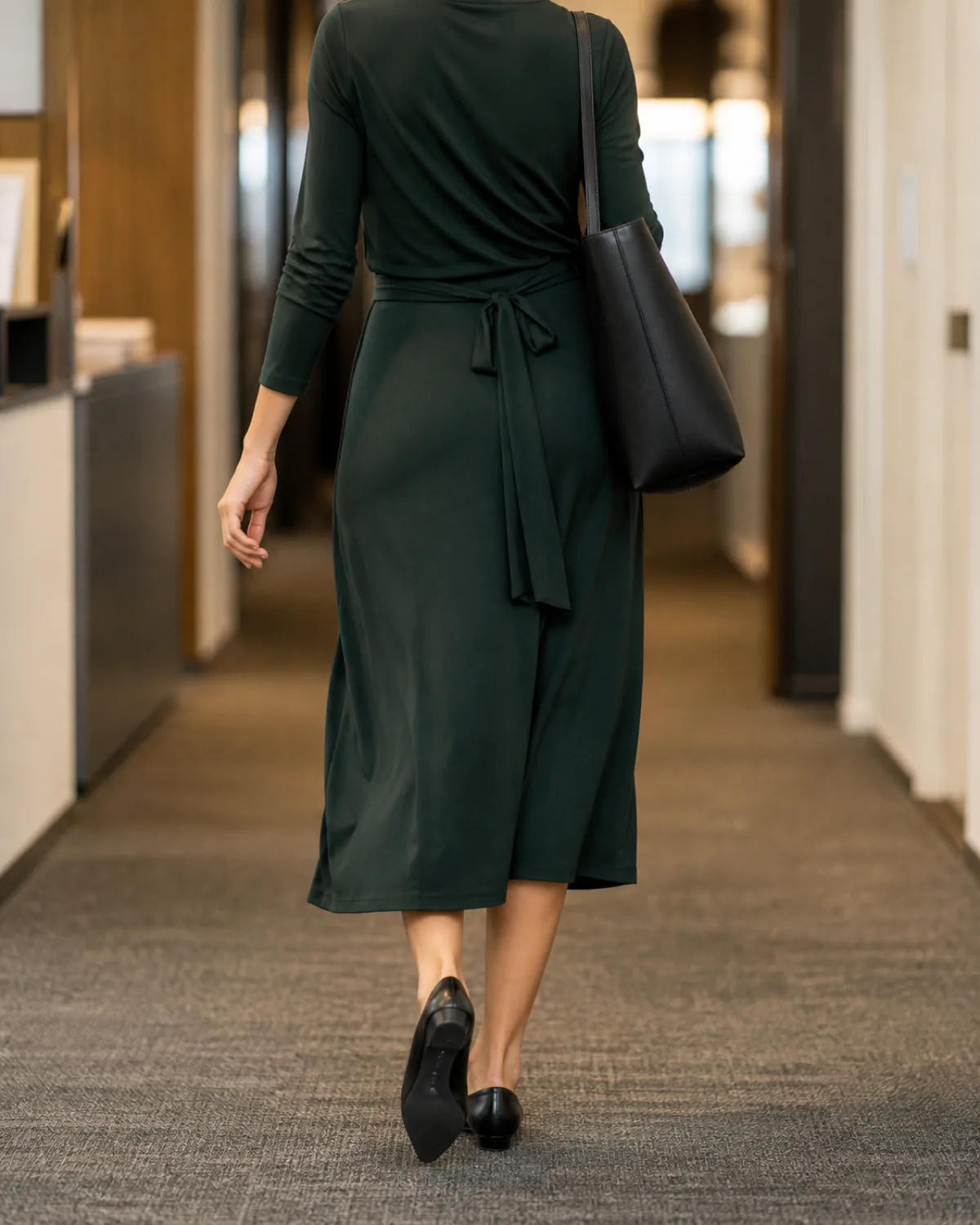 A woman walking away down an office corridor in a dark-green matte jersey midi wrap dress with a black tote bag on her shoulder