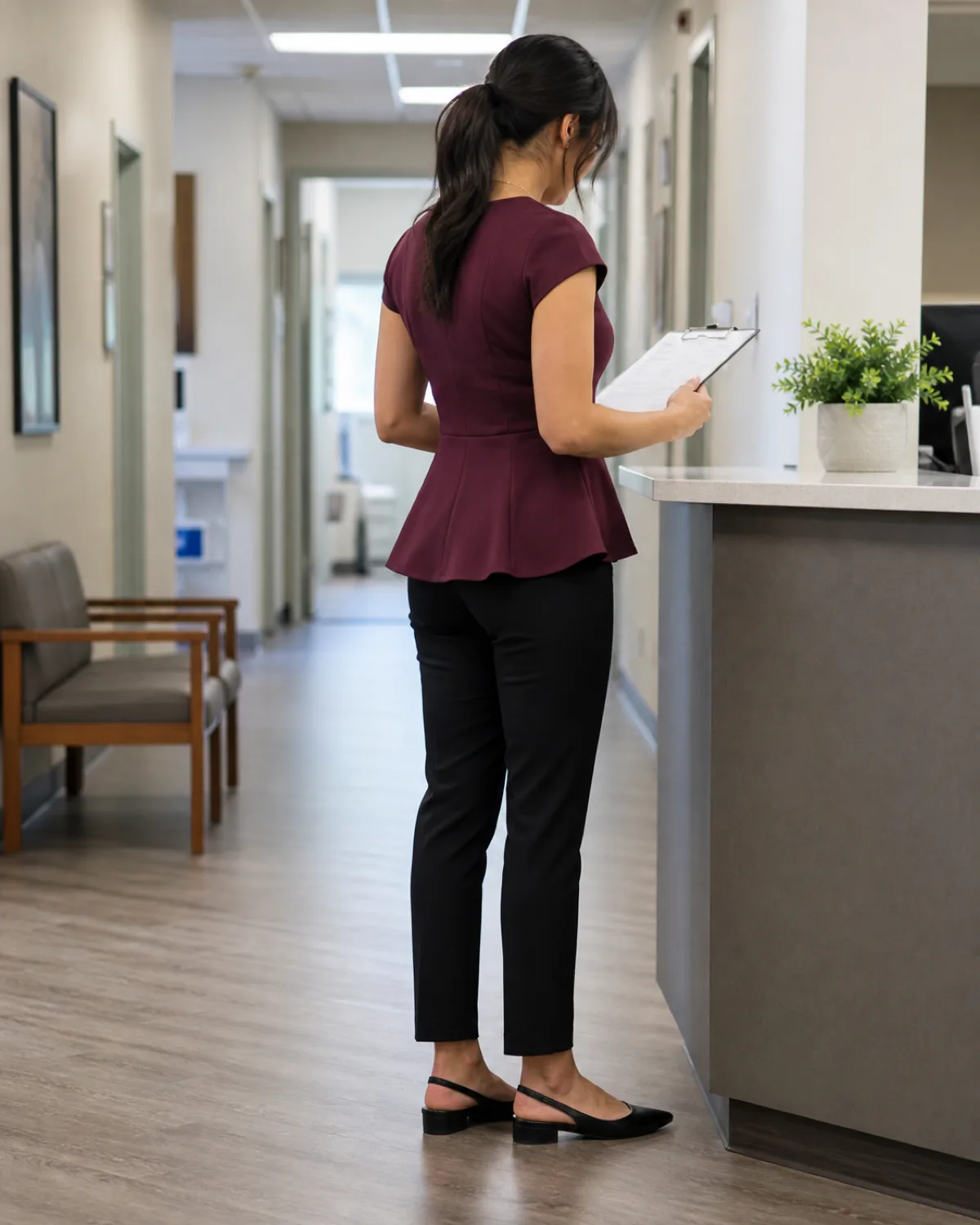 A woman in a burgundy peplum top and black ponte trousers standing at a medical office reception counter, looking down at a clipboard in a back-three-quarter view