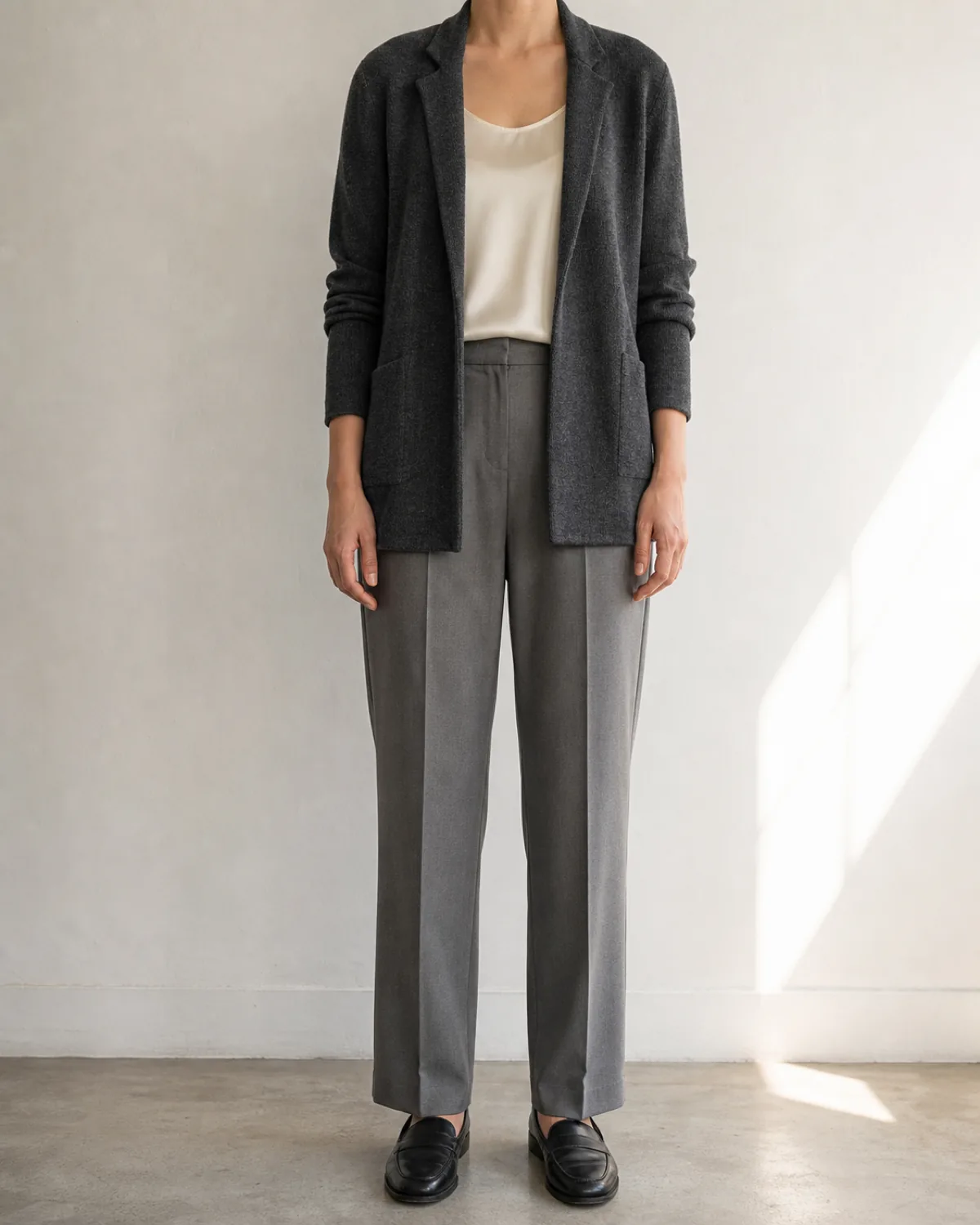 A woman standing straight-on in a dark charcoal knit blazer over a cream silk shell and mid-grey trousers with black loafers