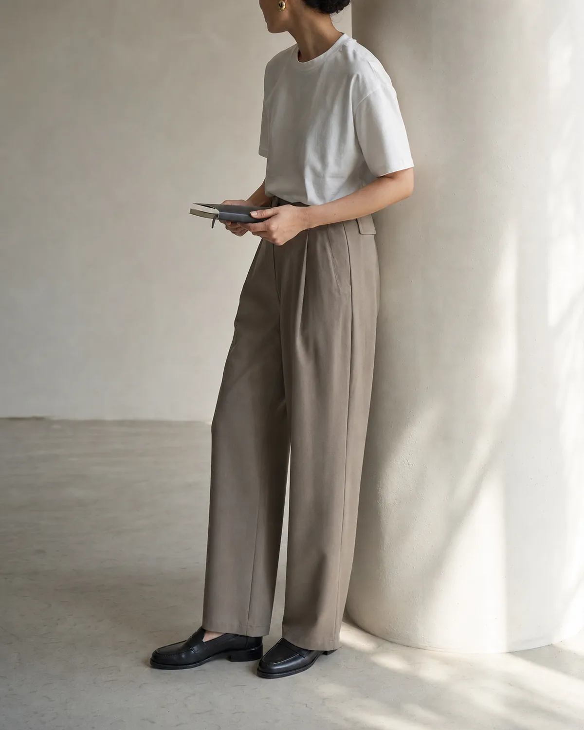 A woman in a three-quarter back view leaning against a plain column, wearing a white heavyweight crew-neck t-shirt tucked into taupe relaxed-fit trousers with black loafers and a small gold earring
