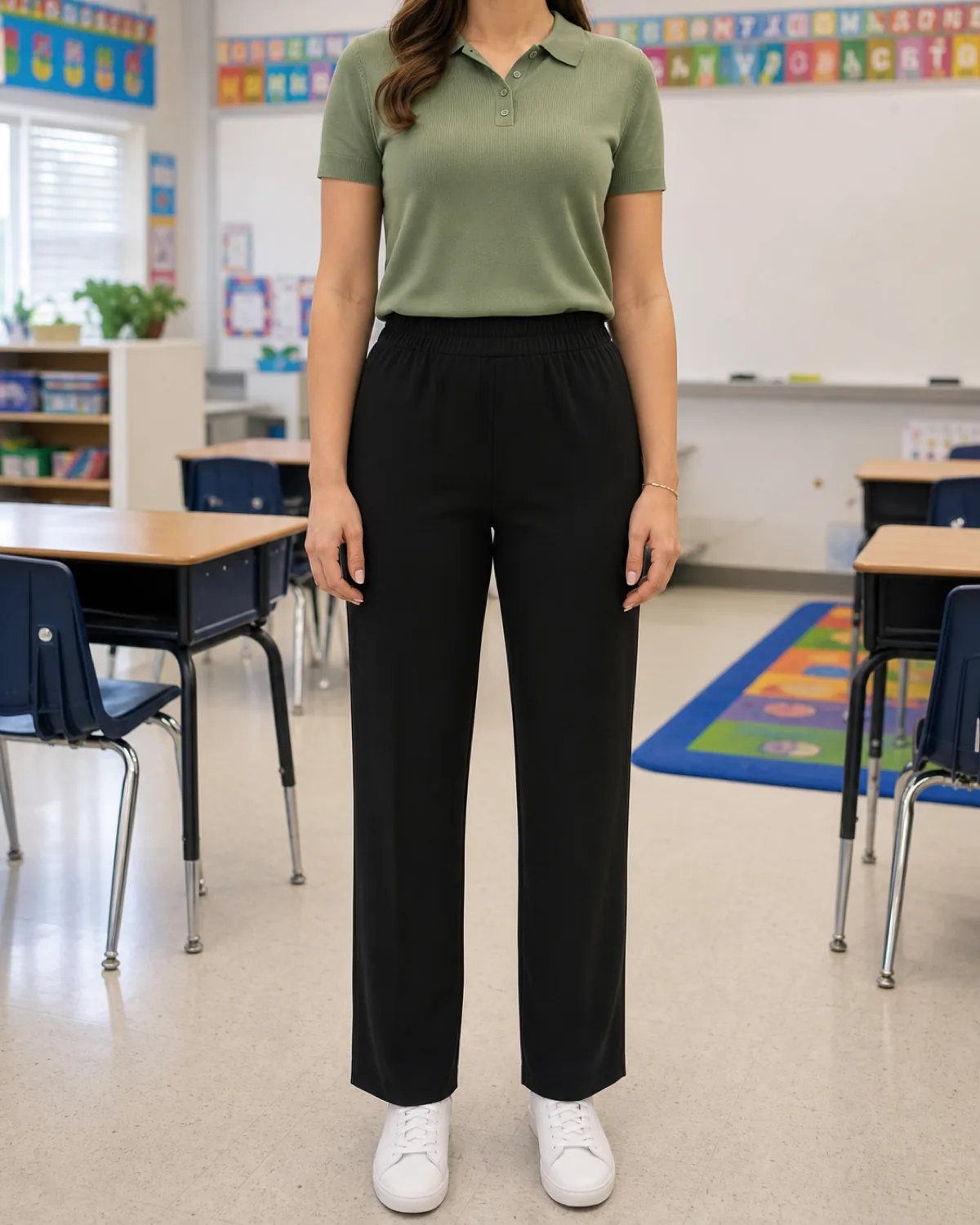 A woman standing straight-on in an elementary school classroom wearing a sage-green knit polo tucked into black pull-on straight-leg trousers with white sneakers, child-height desks and an alphabet border visible behind her