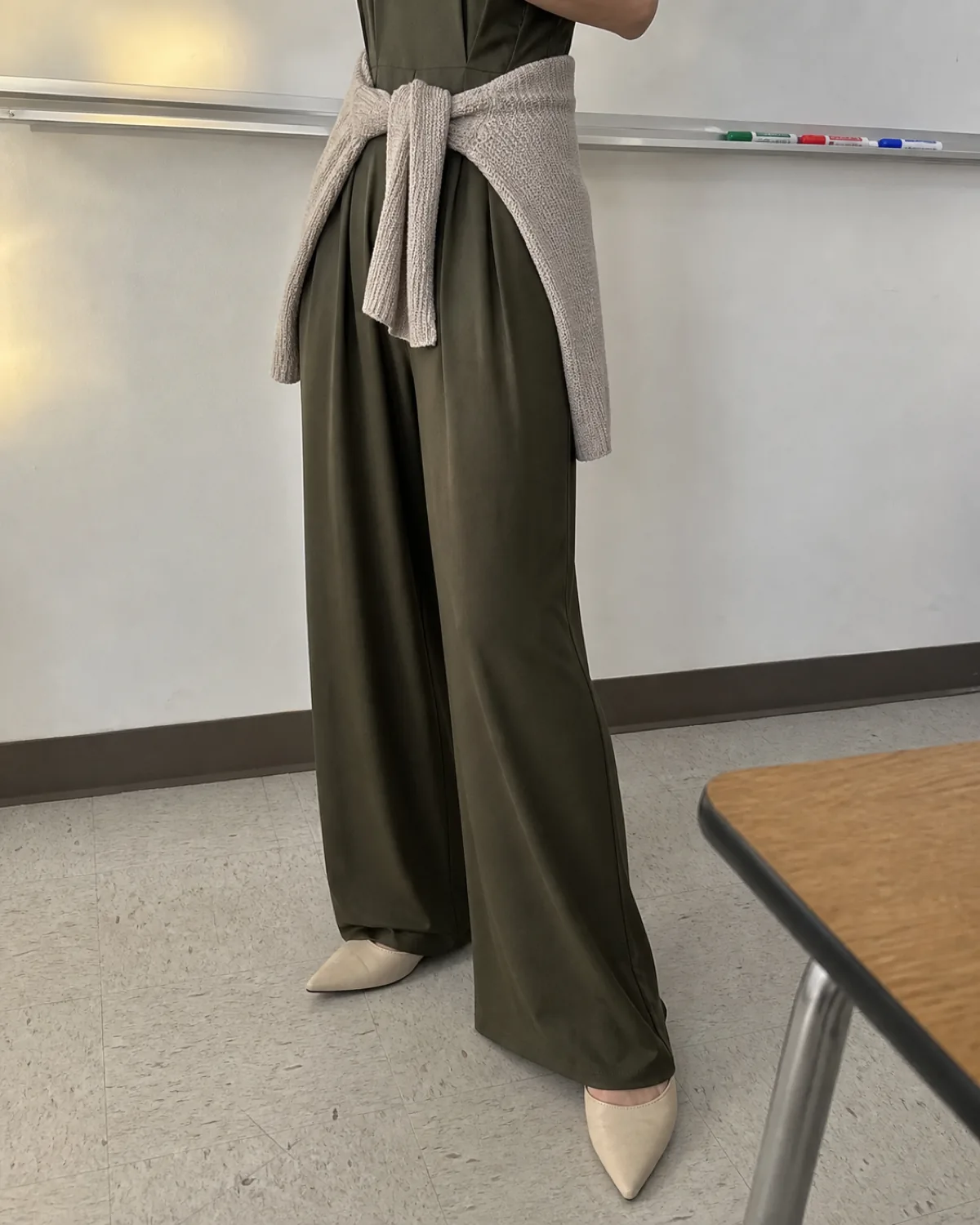 Close crop on the hem and shoes of a woman in a high school classroom wearing a dark olive wide-leg jumpsuit with an oatmeal cardigan tied at the waist and cream pointed-toe flats, whiteboard edge and marker tray visible