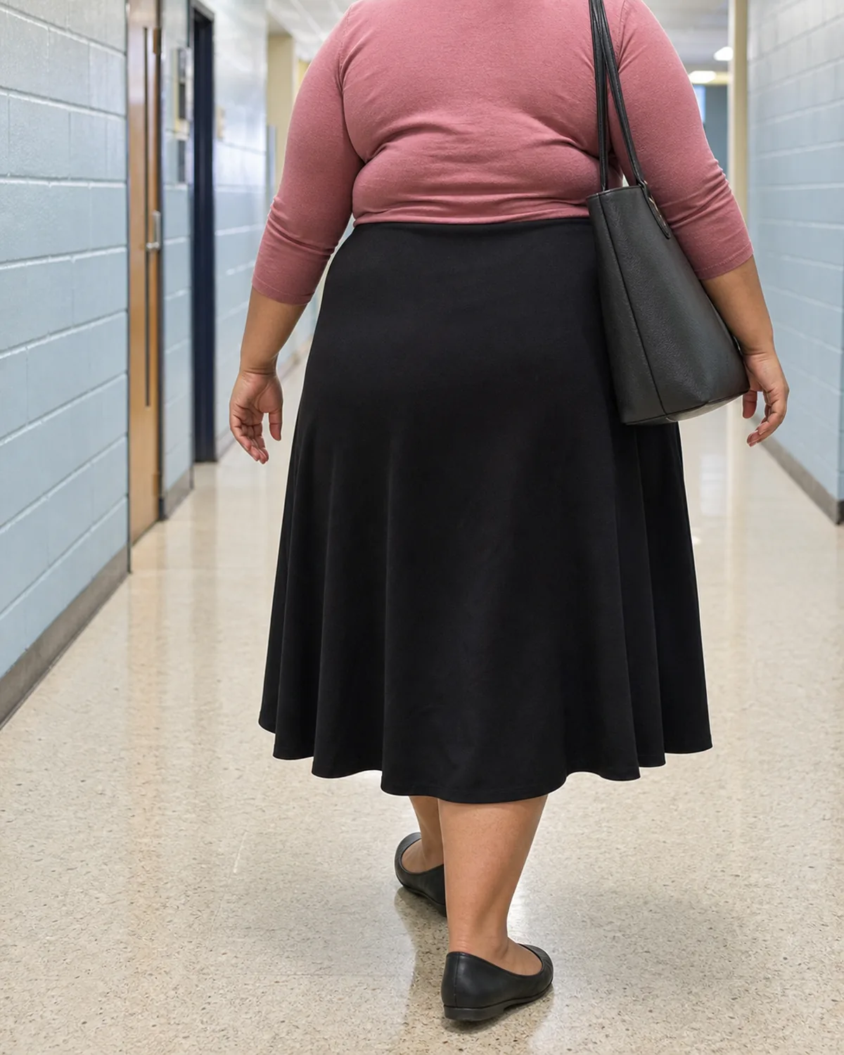 A plus-size woman walking away down a school hallway, wearing a dusty-rose fitted knit top with three-quarter sleeves and a black A-line midi skirt with black closed-toe flats, carrying a dark tote bag