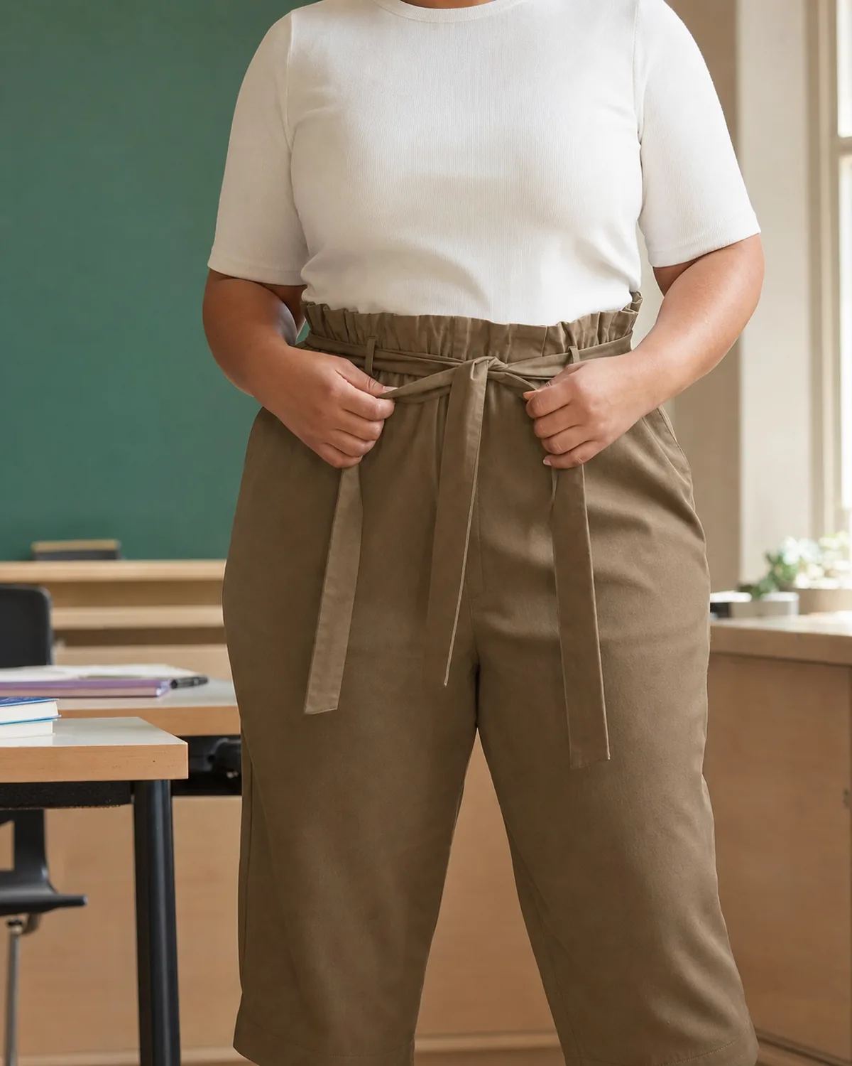 Mid-torso crop of a plus-size woman in a classroom, hands adjusting the gathered waistband of dark khaki paper-bag waist trousers with a self-fabric belt tie, wearing a white short-sleeve crew-neck top